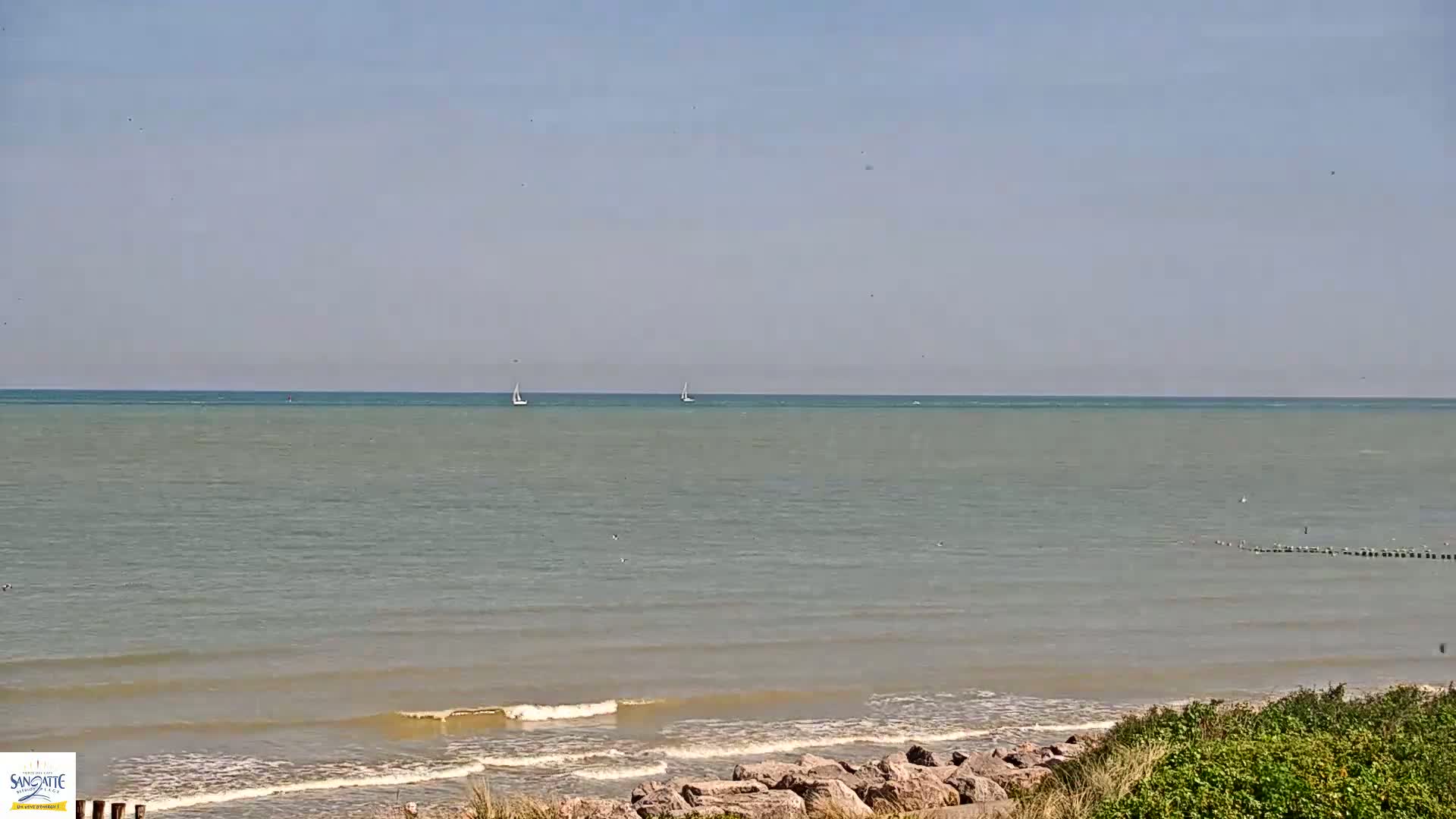 A calm, light-green sea stretches to the horizon under a clear, pale-blue sky, with two sailboats visible in the distance and small waves lapping a rocky shoreline.