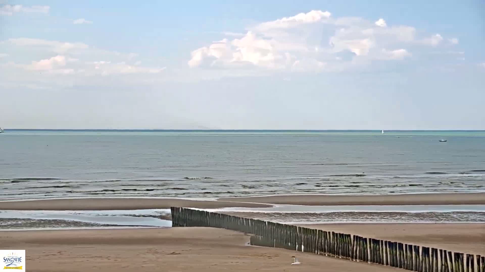A sandy beach with a wooden fence and calm ocean under a partly cloudy sky.