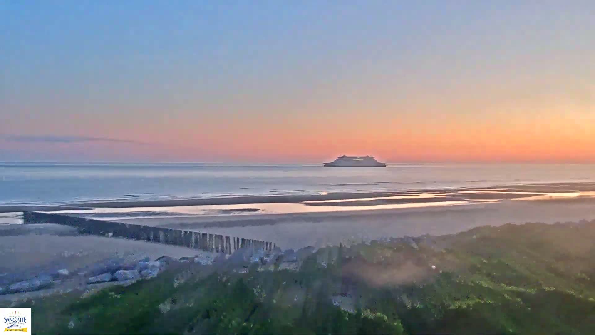 A large ferry is visible on the ocean at sunset, with a sandy beach and vegetation in the foreground.