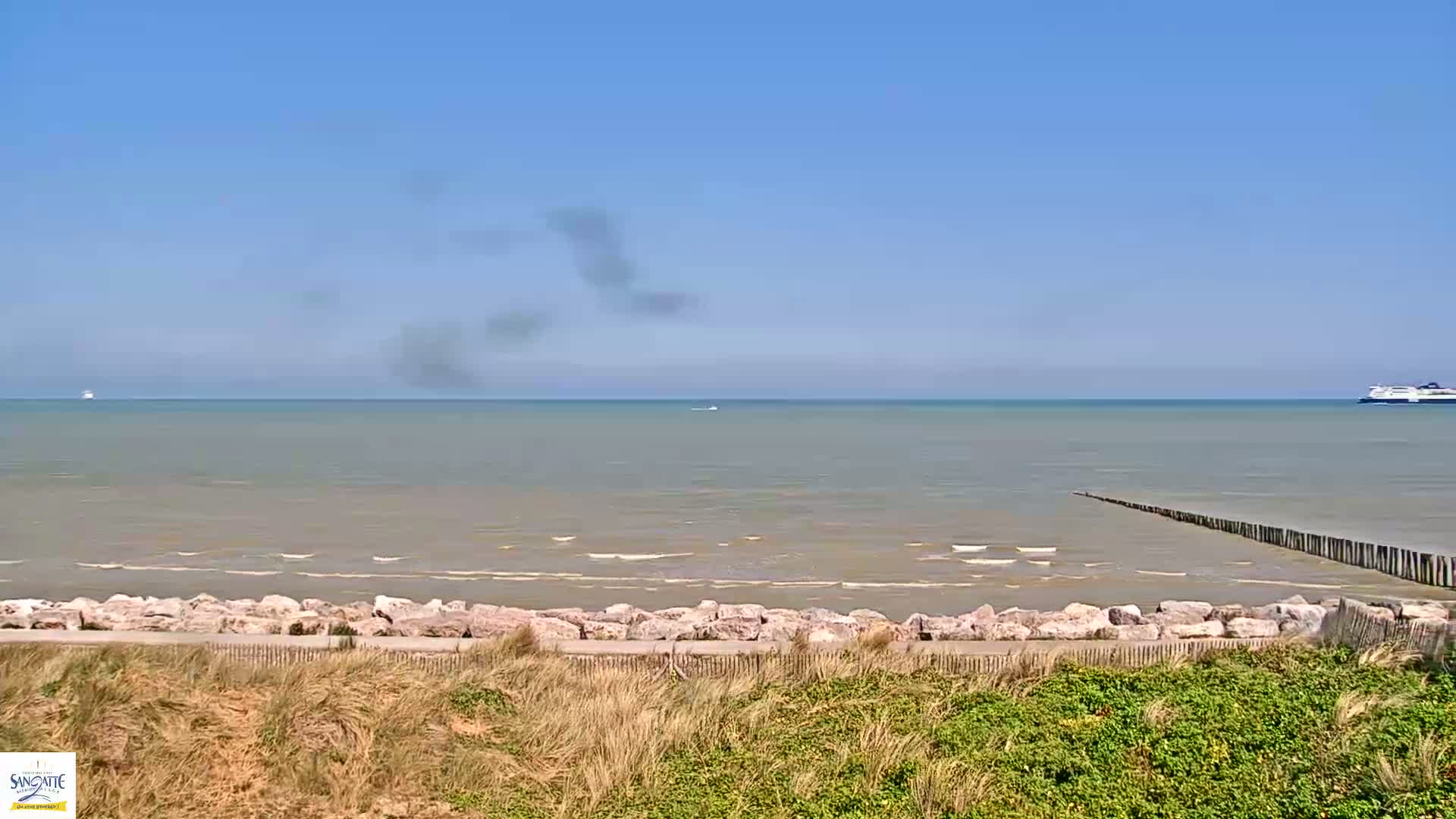 A calm sea with a distant ferry and a breakwater is viewed from a grassy dune under a clear blue sky.