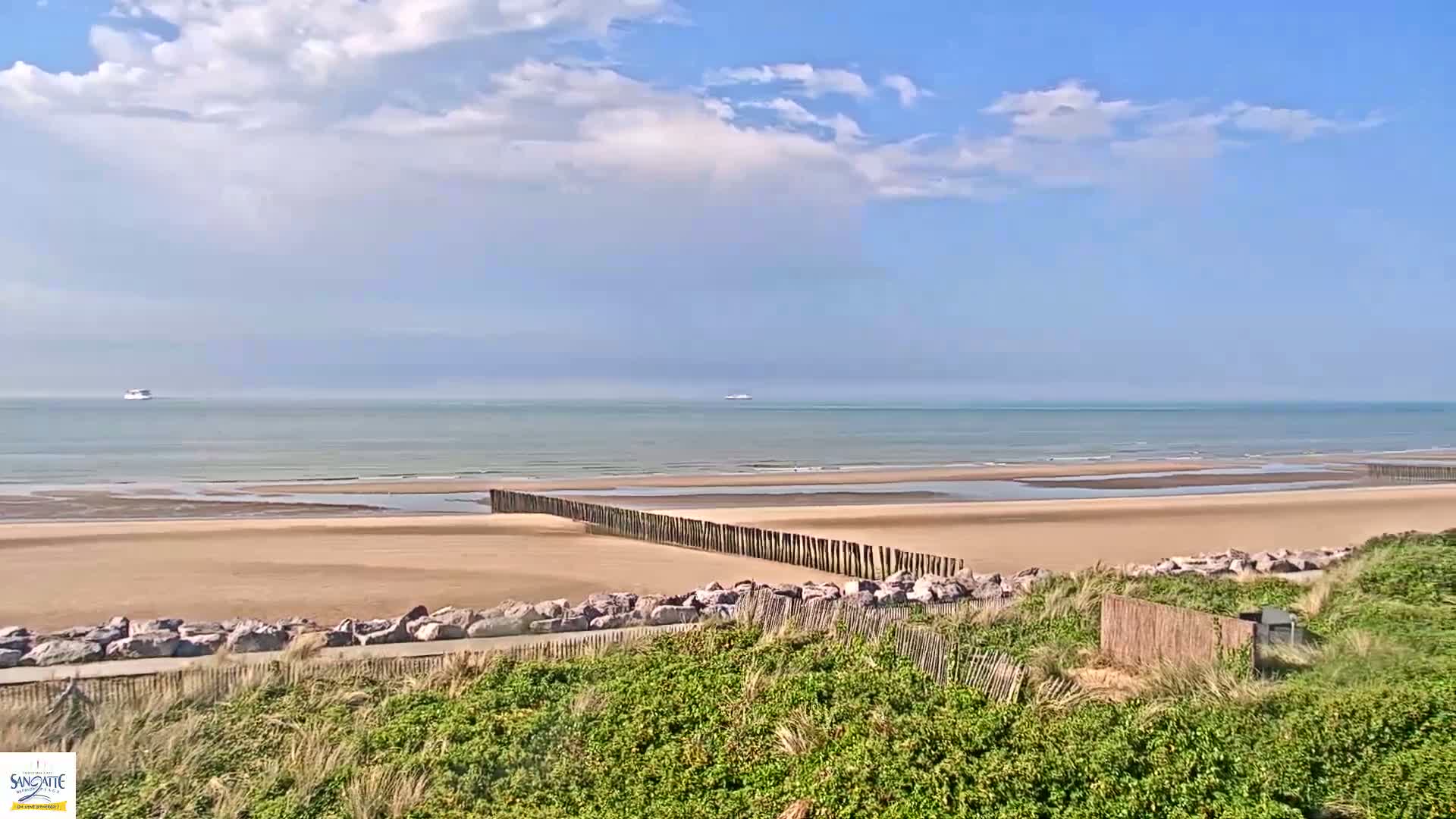 A sandy beach stretches to a calm ocean under a partly cloudy blue sky, with a wooden barrier partially separating the beach from a grassy dune area.