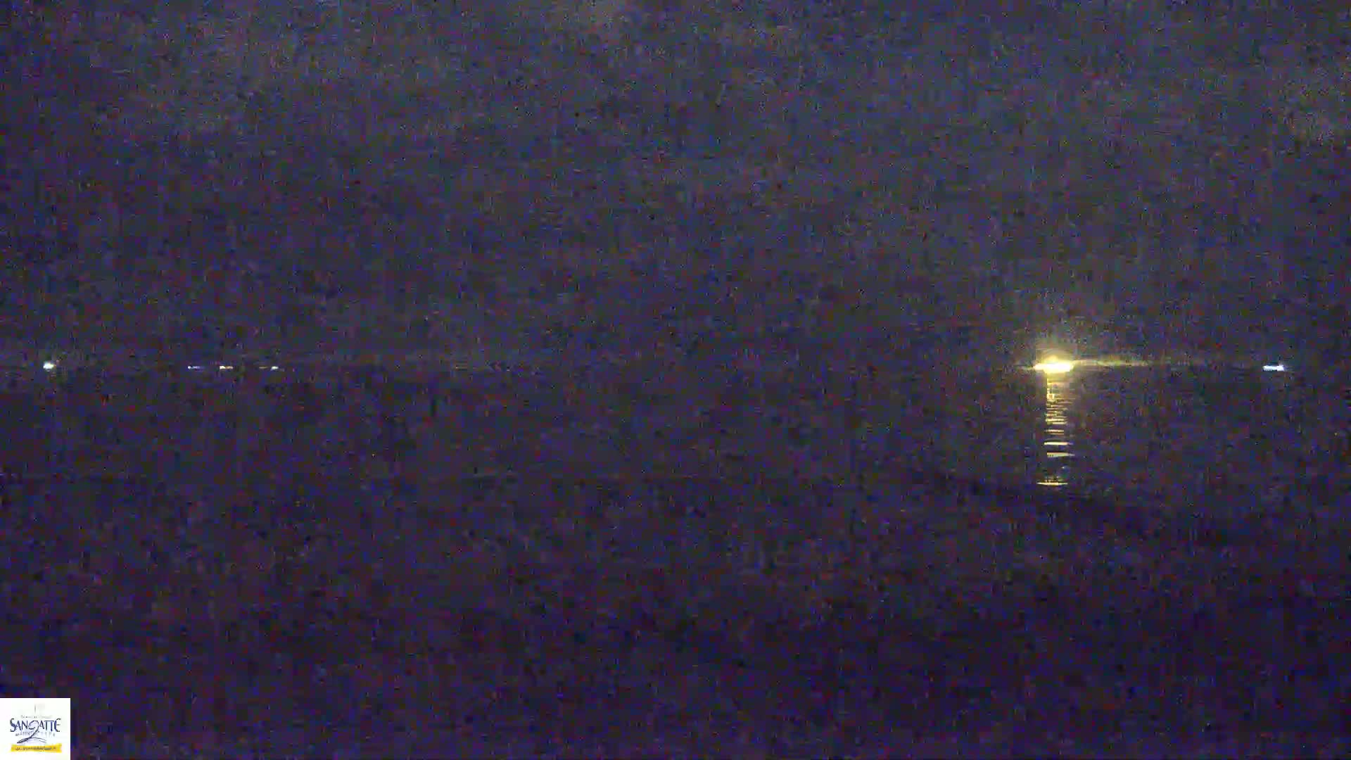 A nighttime view of a dark body of water reflecting a distant light source under a cloudy sky.