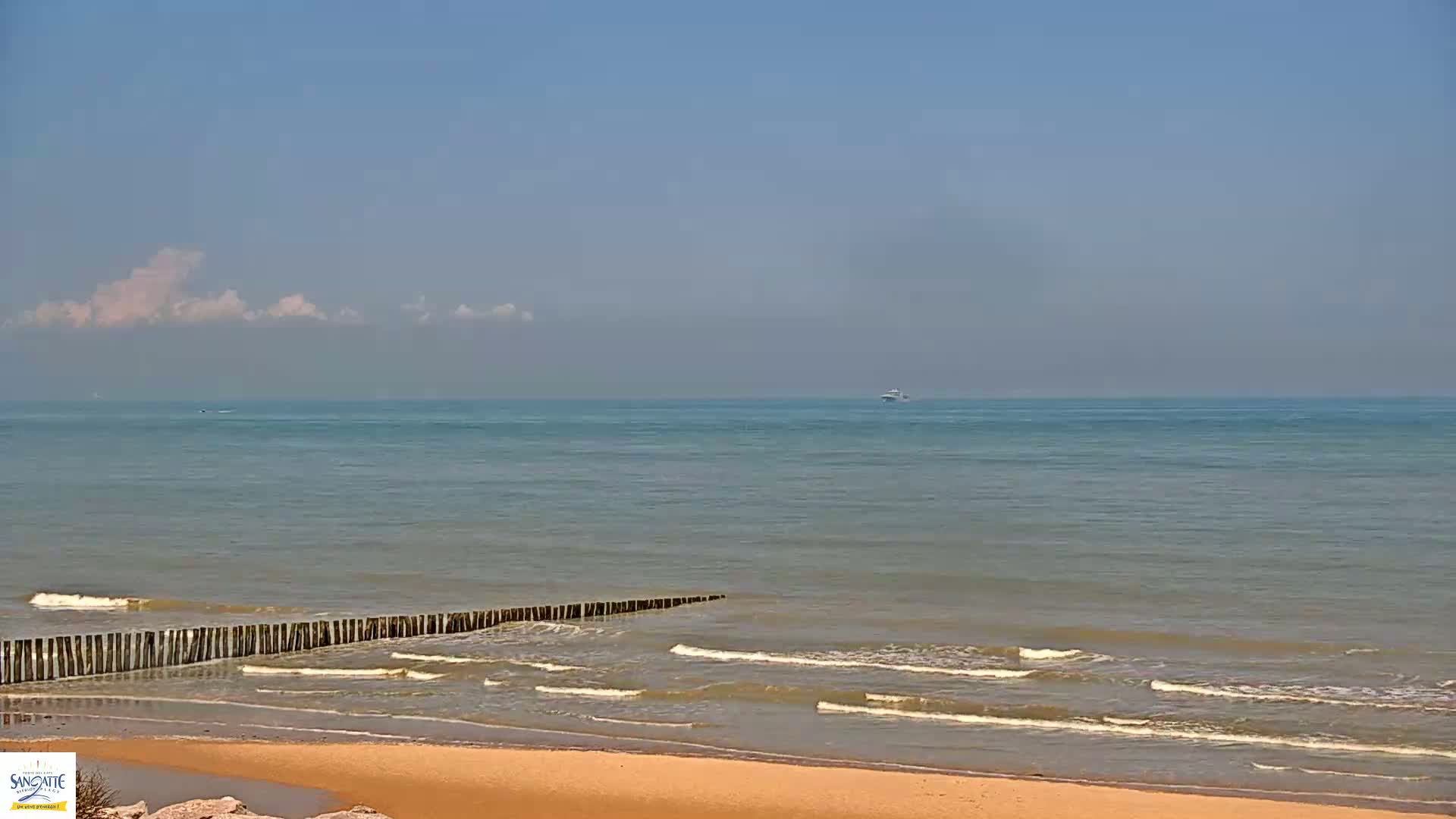 A sandy beach with gentle waves lapping against a low wooden breakwater under a mostly clear sky with a few wispy clouds.