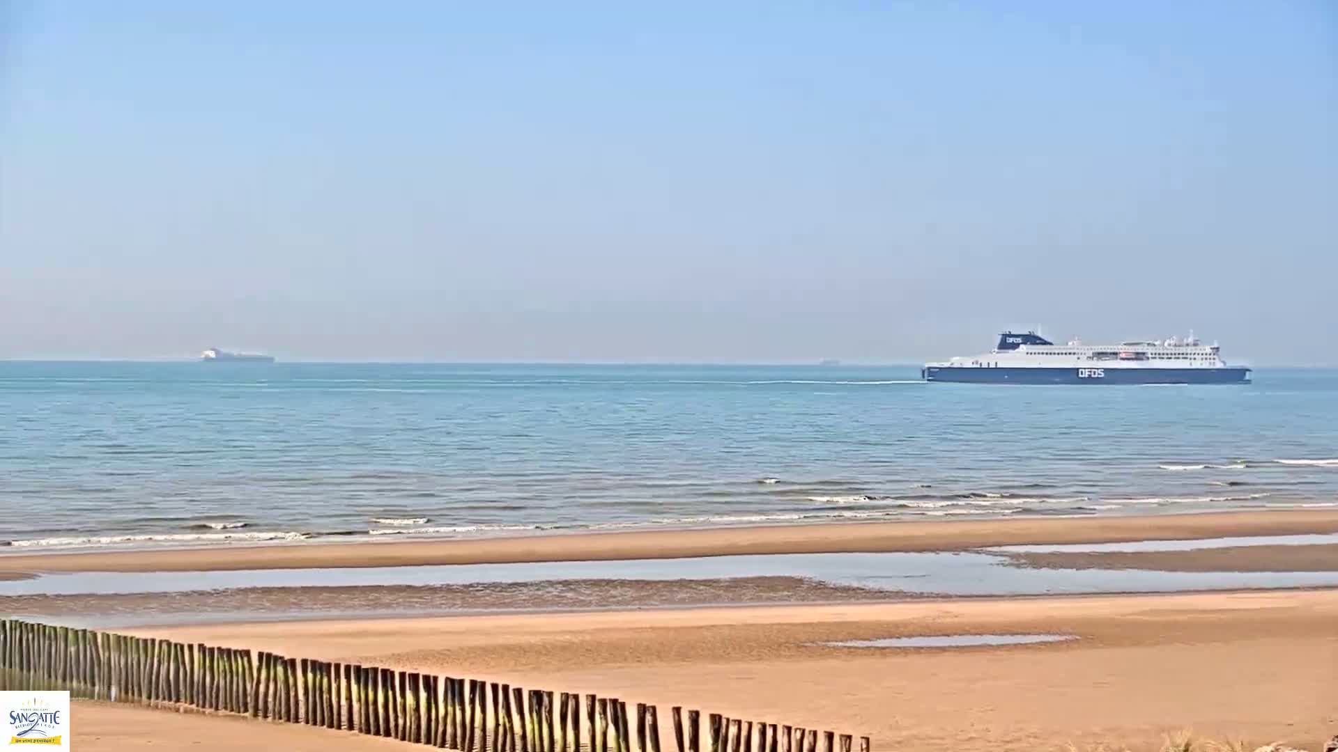 A sandy beach with a wooden fence in the foreground, and two large ships visible on a calm, clear ocean in the background.