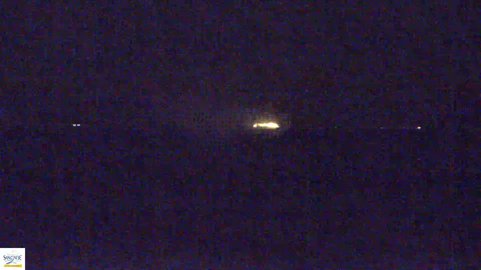 A blurry, nighttime image shows a bright light in the distance, possibly a distant vehicle's headlights, with faint lights visible further away.