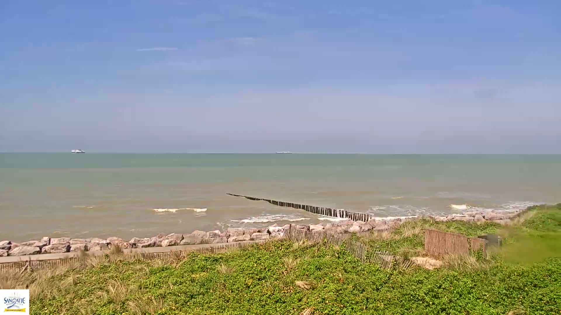 A calm, somewhat cloudy day at the beach shows a rocky shore with low-lying vegetation, a breakwater extending into the sea, and two distant ships on a greenish-grey ocean.