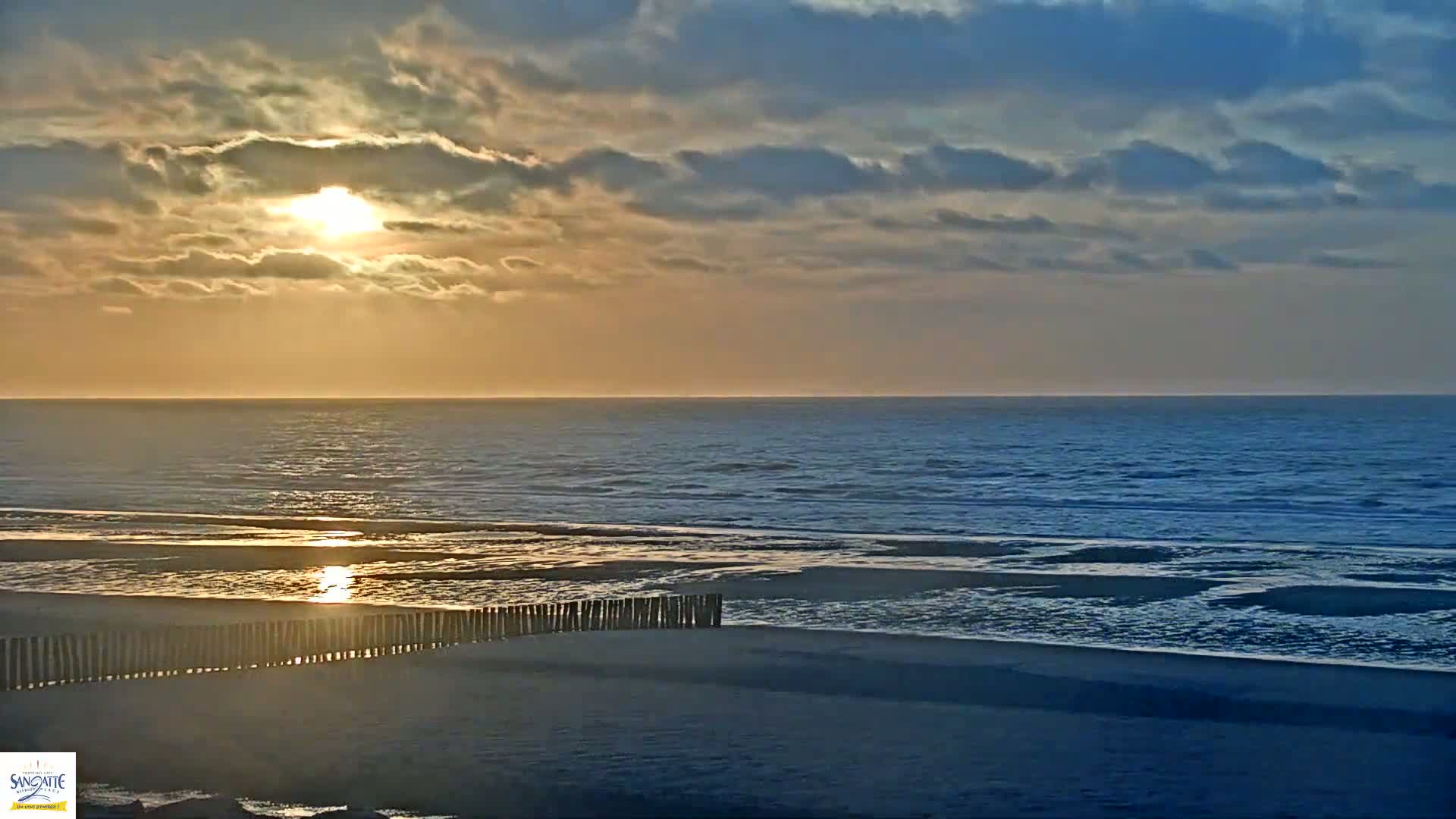 A partly cloudy sunrise casts a golden glow over a calm ocean beach with a low wooden fence separating the sand from the water.