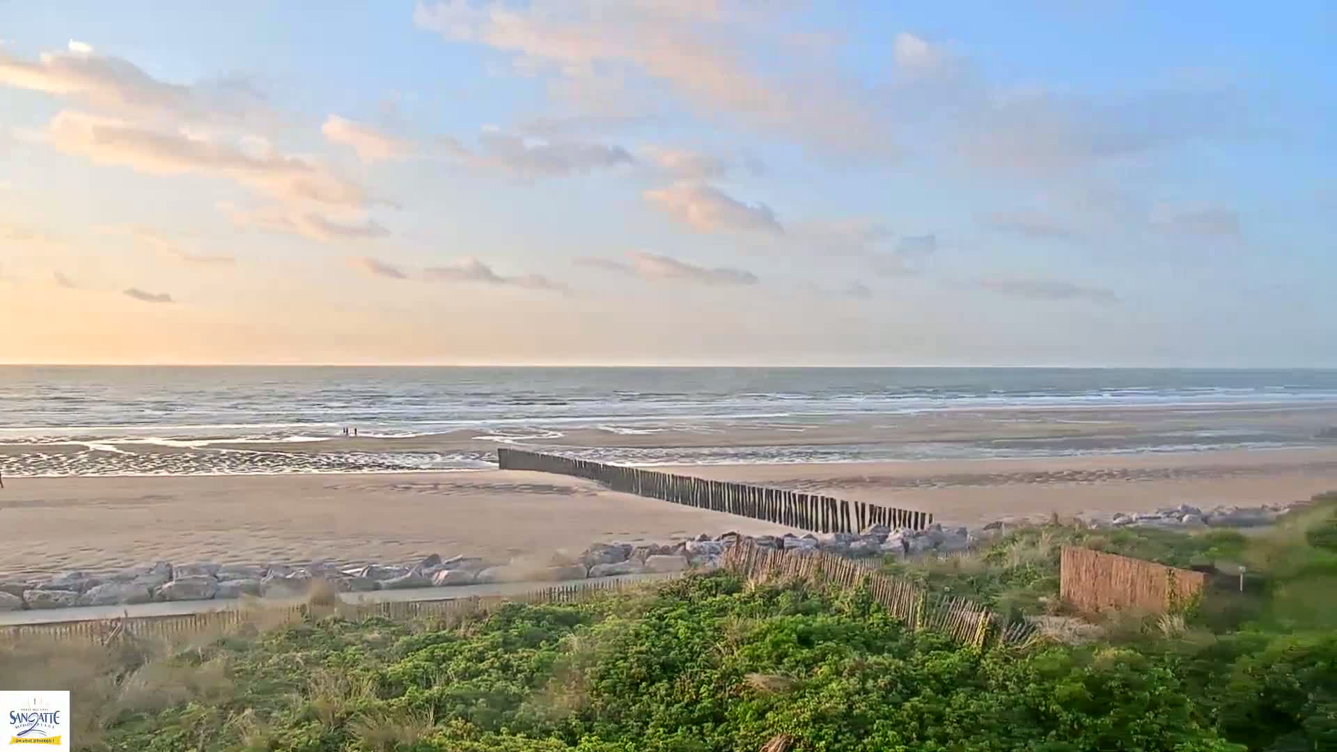 A sandy beach with a wooden post fence extends into the ocean under a partly cloudy, pale sky.