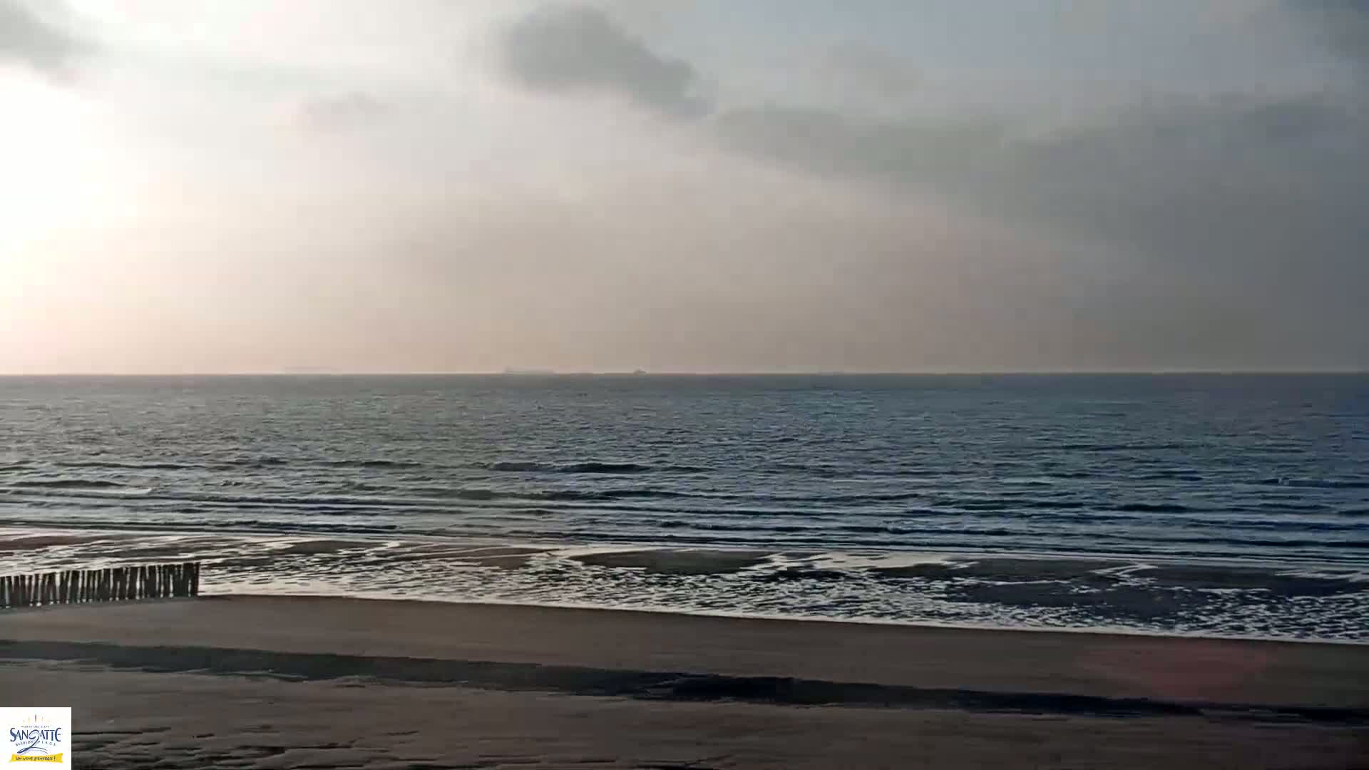 A sandy beach with gentle waves rolls onto the shore under a mostly cloudy, light sky.