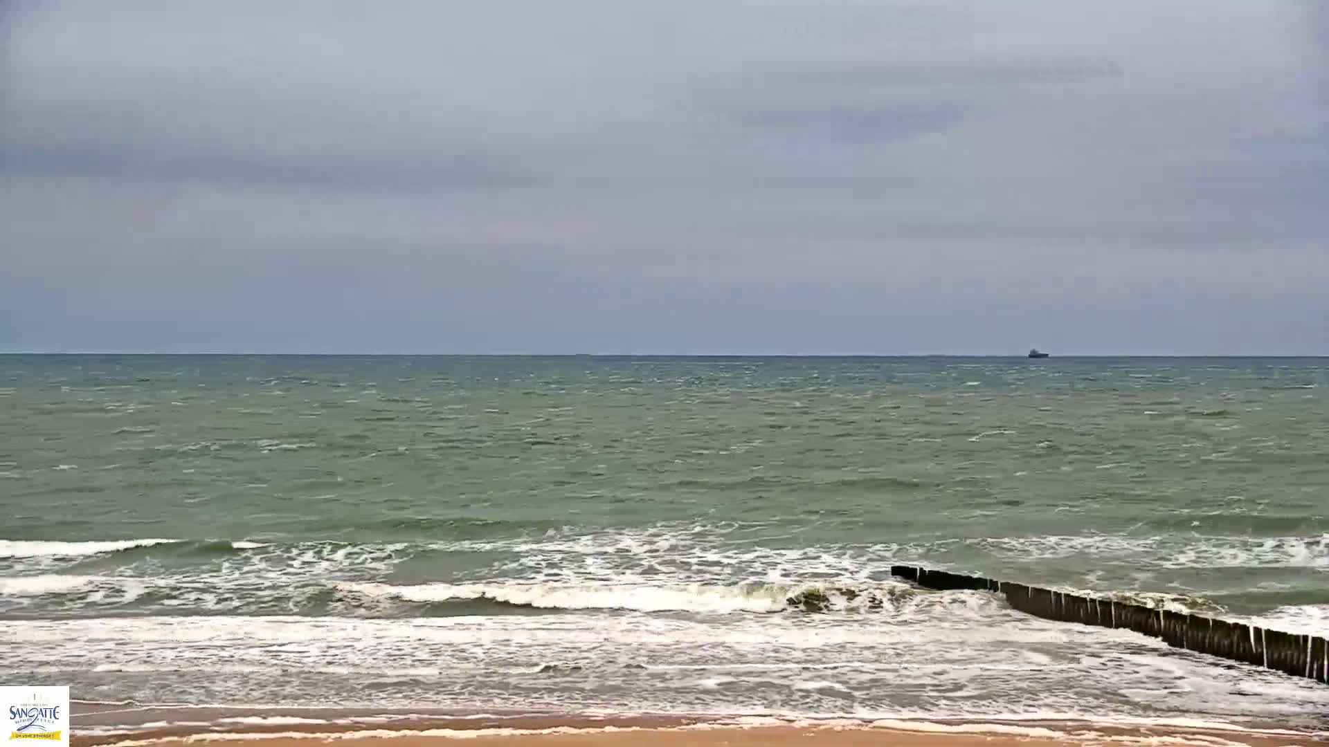 A sandy beach with moderate waves crashing against a breakwater under an overcast sky.