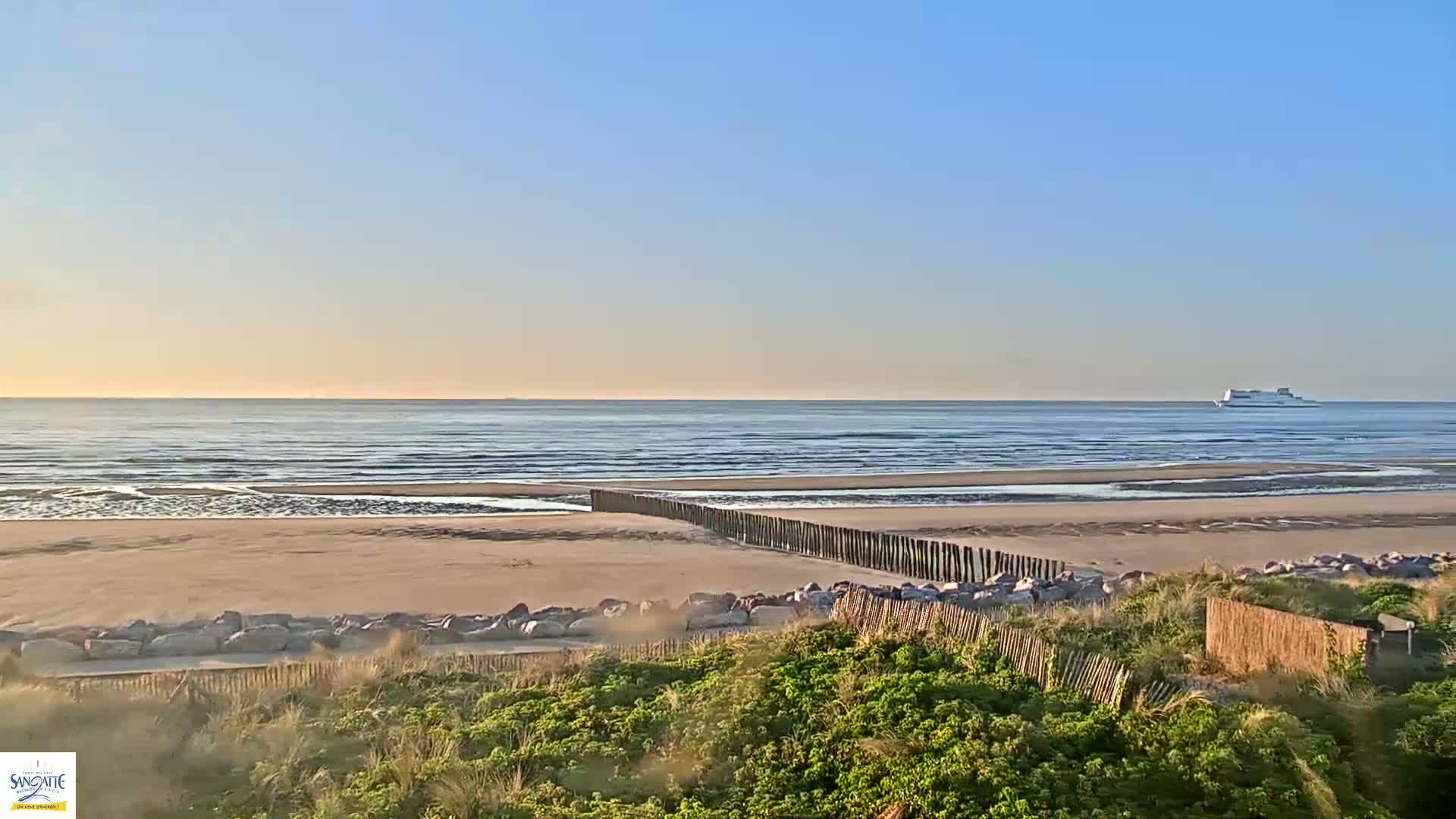 A sandy beach with a wooden fence and a distant ferry is visible under a clear, bright sky.