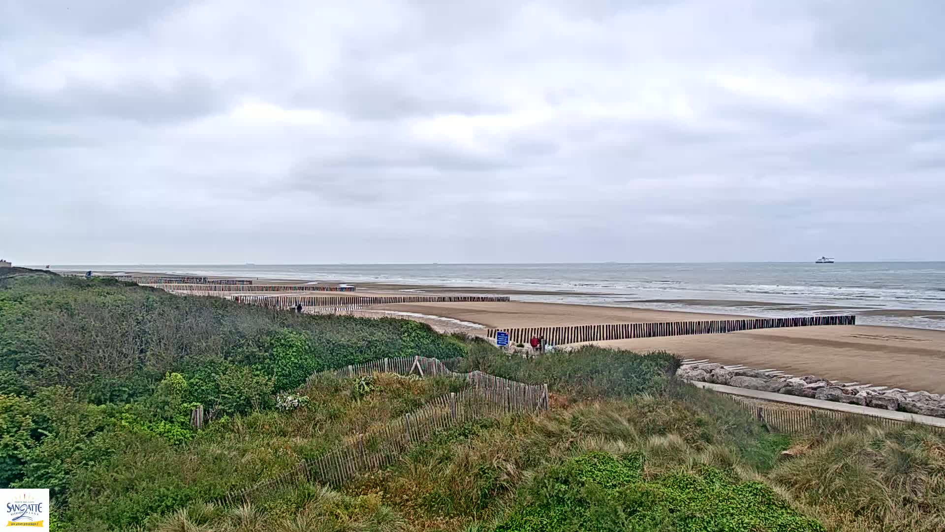 A sandy beach with a long line of wooden posts along the water's edge is partially visible from behind a dune covered with green vegetation under a cloudy sky.