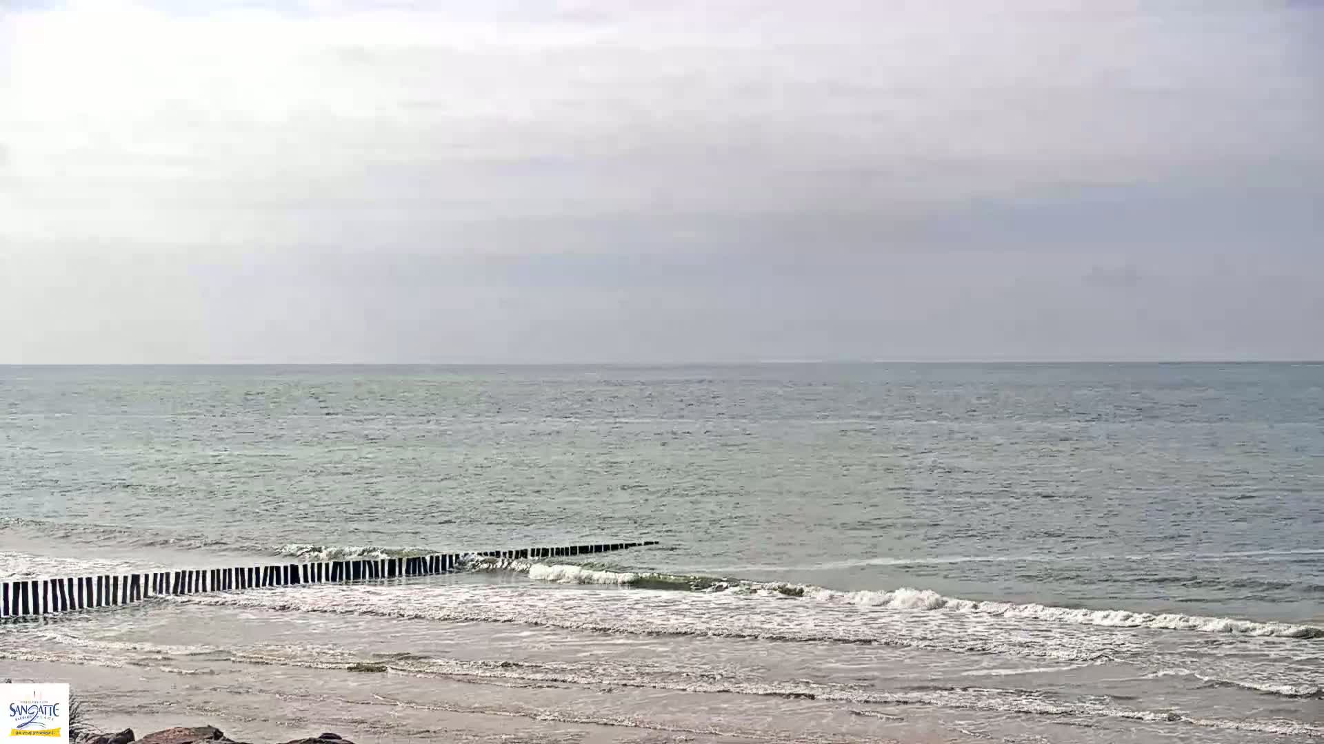 A calm sea with small waves rolling onto a sandy beach protected by a low, dark wooden breakwater under a cloudy sky.