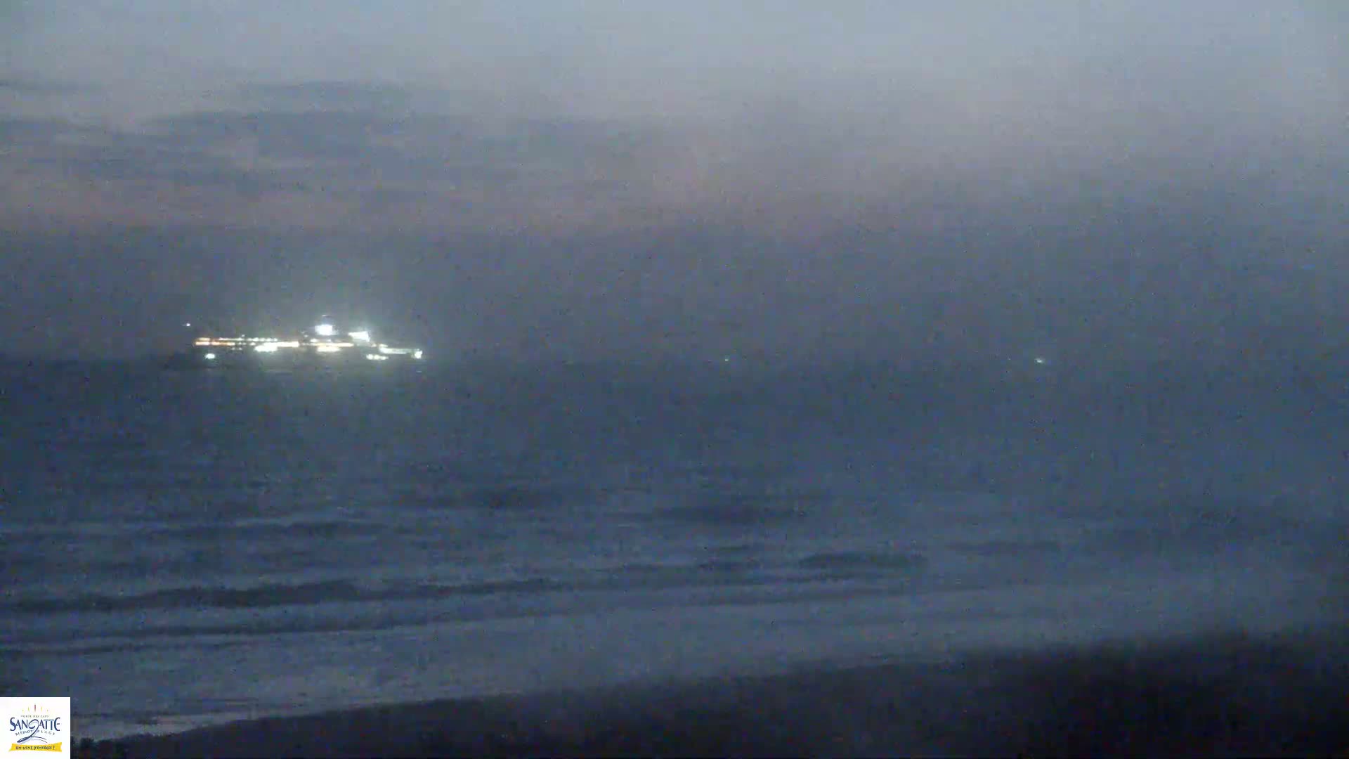 A brightly lit ship is visible on the horizon over a dark, wavy ocean under a hazy, dusky sky.