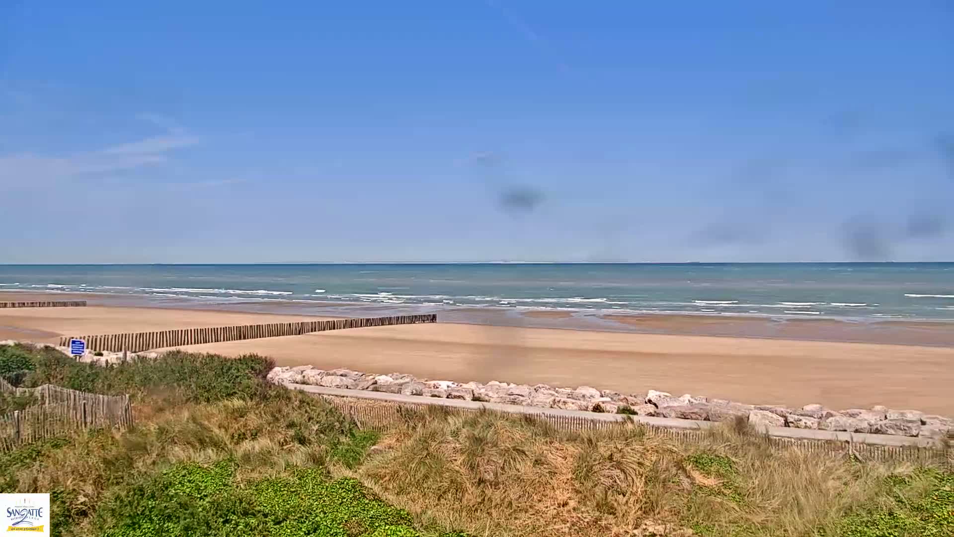 A sandy beach with calm ocean waves under a clear blue sky is viewed from a dune overlooking a low rock wall.