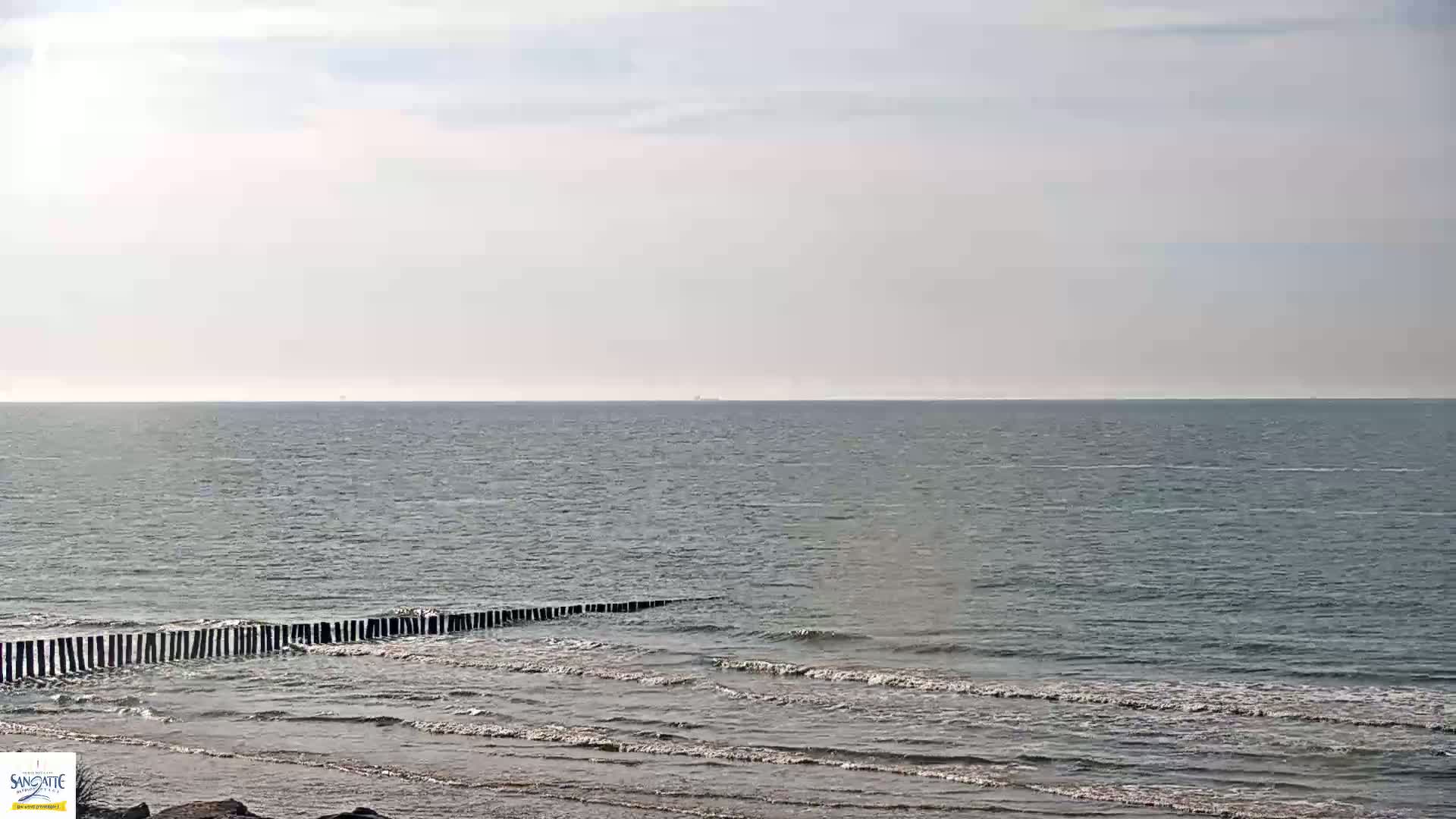 A calm sea with small waves laps a sandy shore protected by a low, dark wooden breakwater under a pale, overcast sky.