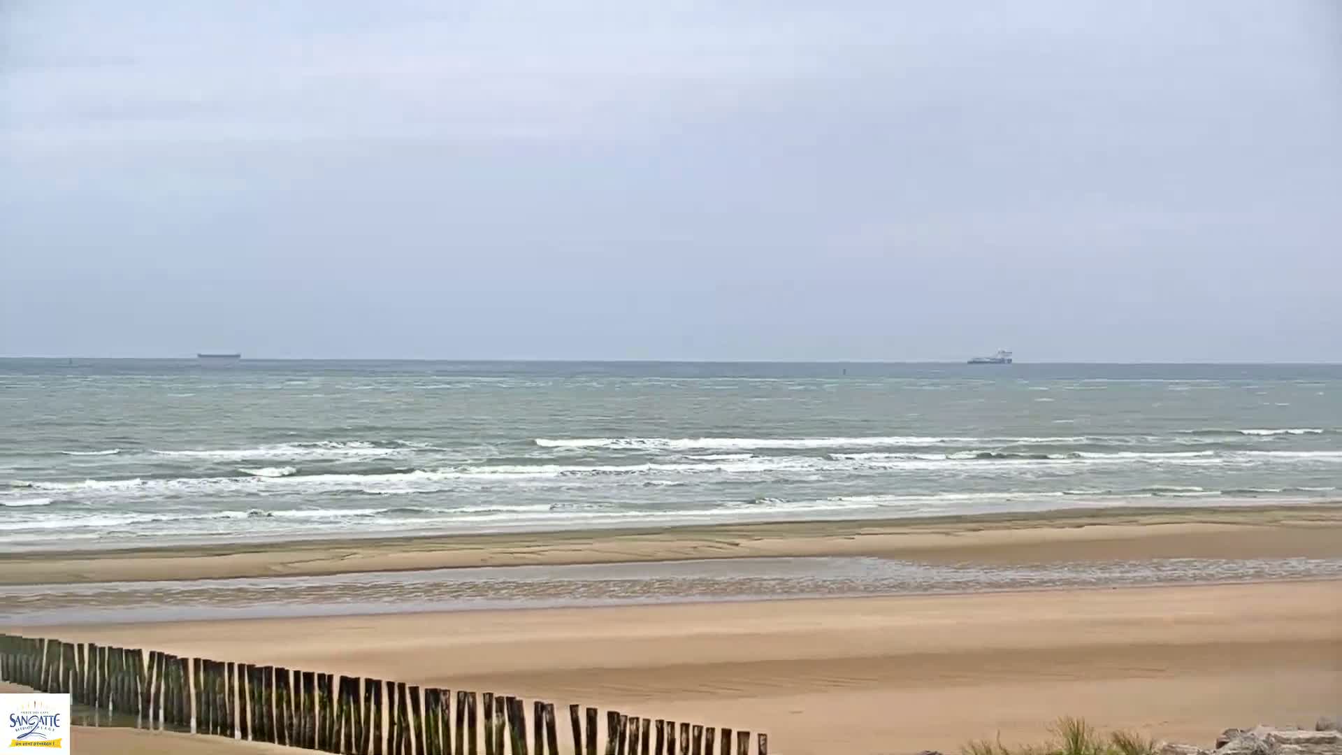 A wide sandy beach with a wooden fence in the foreground and a calm sea with small waves under an overcast sky, showing two ships in the distance.