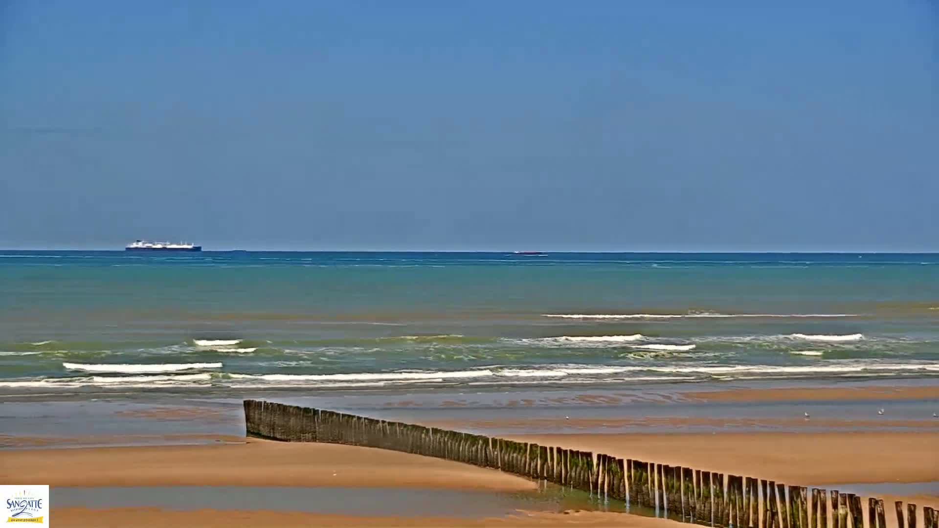 A sandy beach with small waves and a distant ship is visible under a clear blue sky.