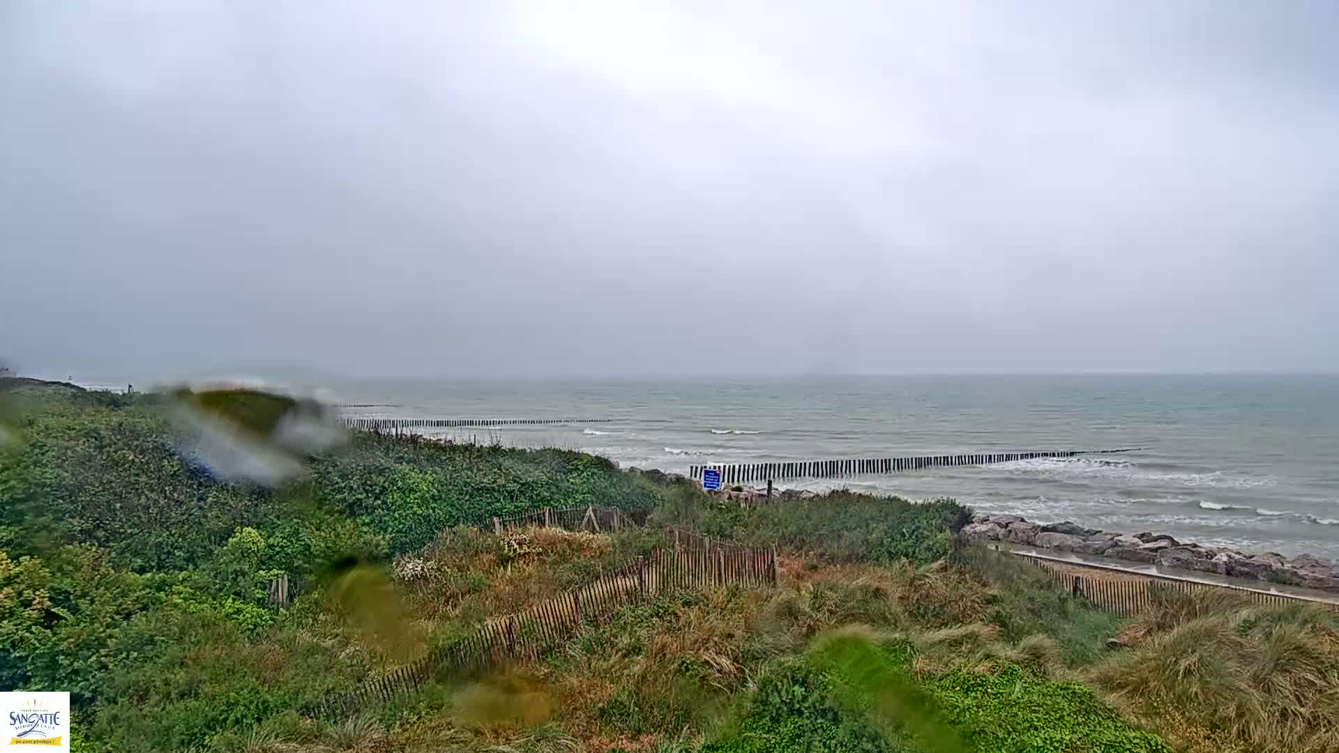 Rain falls on a grassy coastal dune overlooking a choppy sea partially protected by a row of wooden breakwaters.