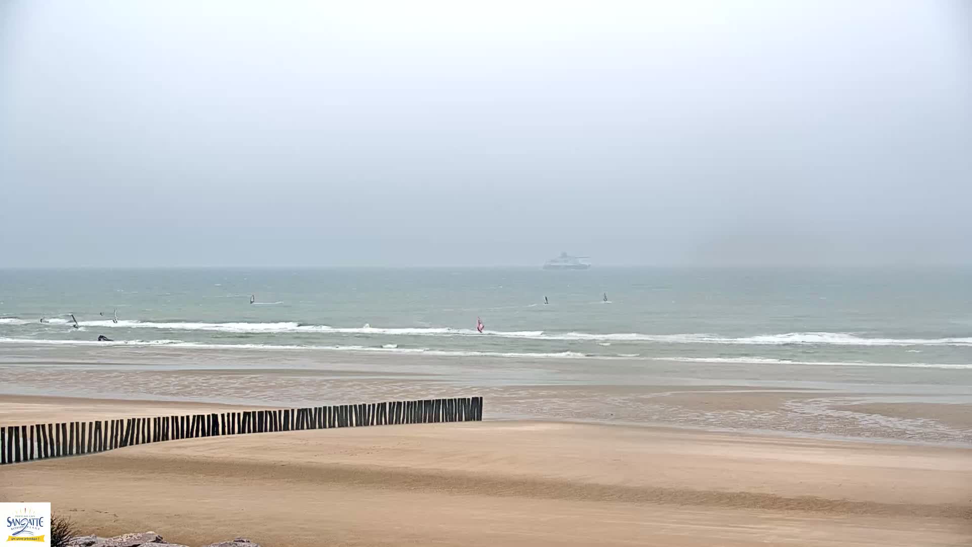 A sandy beach with a low wooden fence separates the sand from a somewhat choppy ocean where several windsurfers are visible on a foggy day.