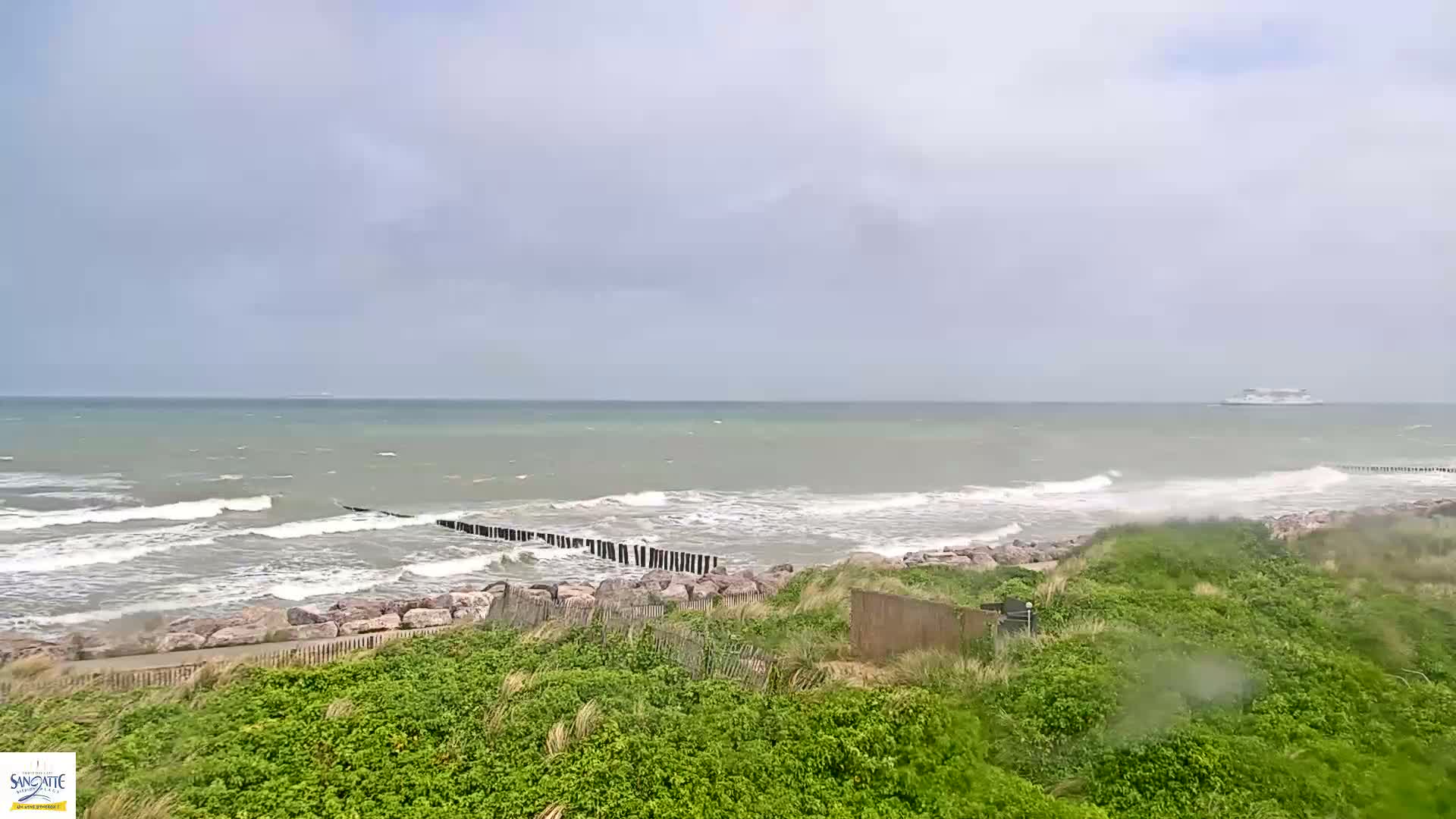 A ferry is visible on the horizon of a choppy sea under an overcast sky, with waves breaking against a rocky shoreline partially protected by a breakwater.