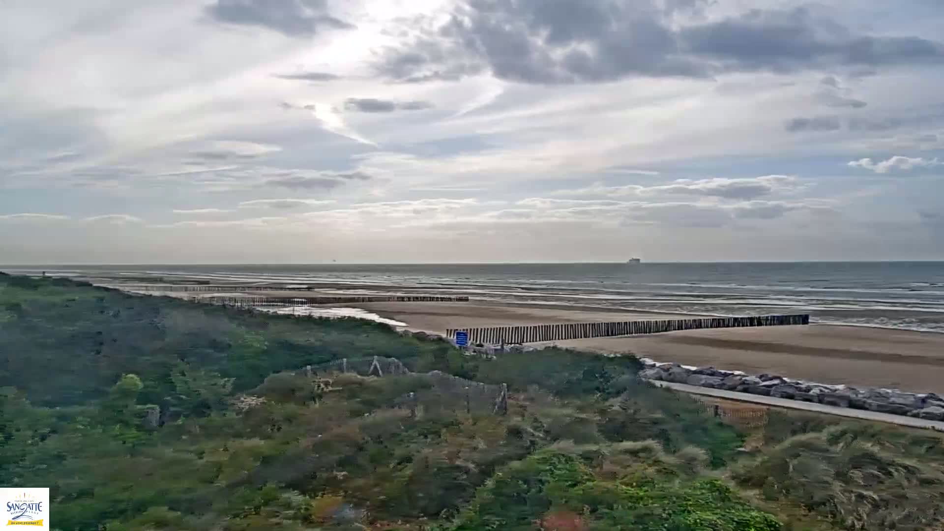 A sandy beach with a wooden barrier stretches along a gently wavy ocean under a partly cloudy sky.