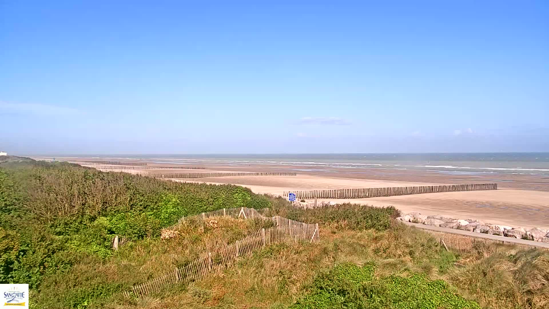 A sandy beach with a wooden fence along the shoreline is visible under a clear blue sky, with gentle waves in the ocean.