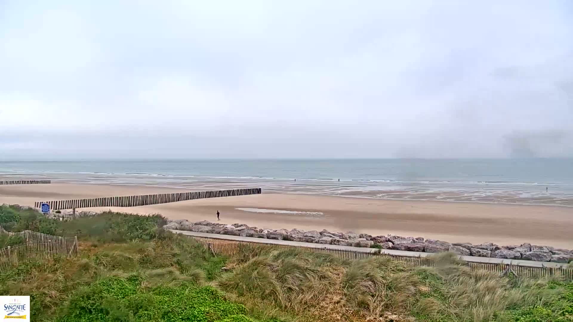 A wide sandy beach stretches to a calm, gray ocean under an overcast sky, with a wooden barrier partially separating the beach from grassy dunes.