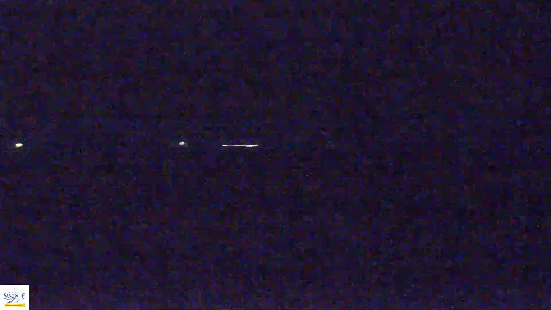 A nighttime image shows a low-lying horizontal light source with two fainter lights on either side, possibly distant lights on a low horizon.