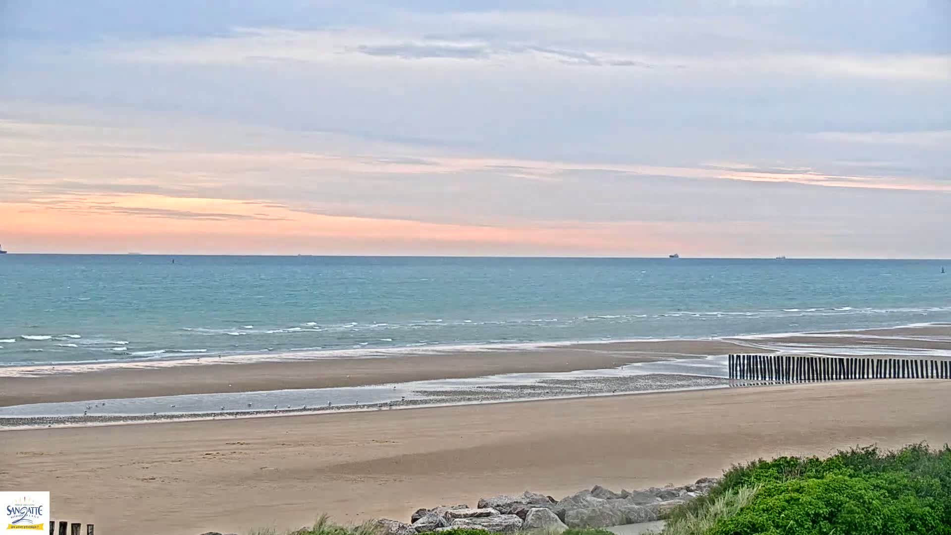 A wide sandy beach with gentle waves under a pale pink and blue sunset sky, featuring a row of wooden posts near the water's edge.