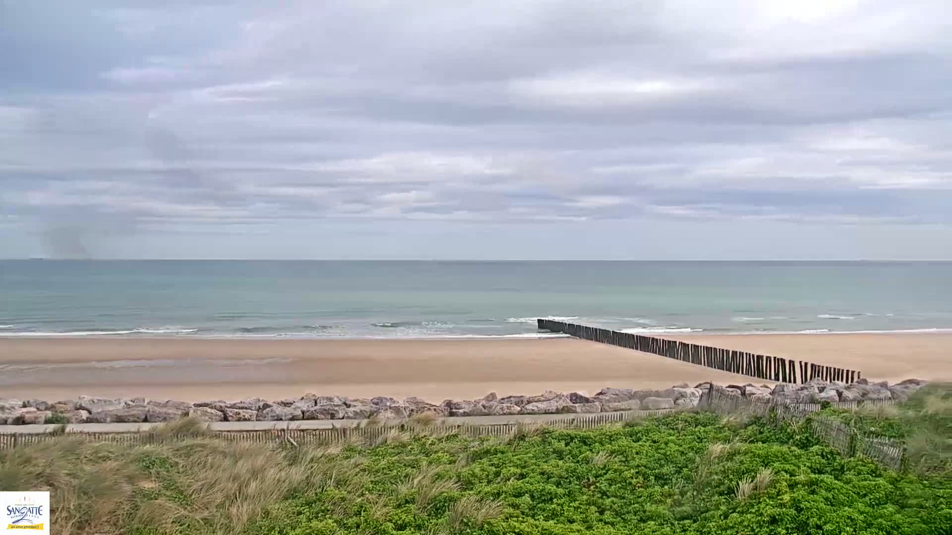 A sandy beach with a low wooden breakwater stretches along a calm, light-green ocean under a cloudy sky.