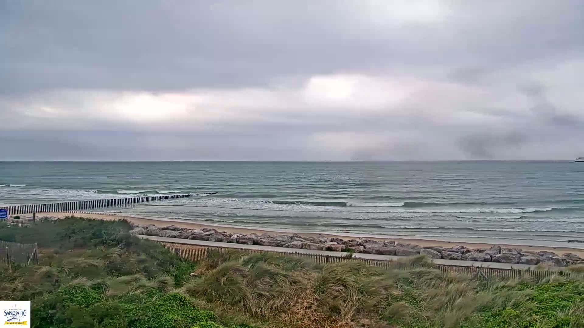 A slightly overcast day shows a beach with moderate waves rolling onto a sandy shore, protected by a rock barrier and grassy dunes.