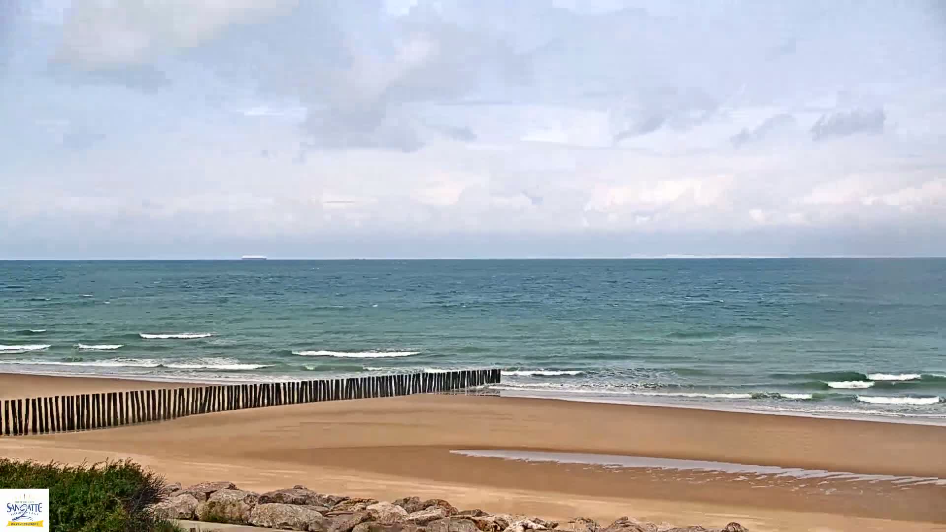 A sandy beach with a low wooden barrier stretches along a gently wavy ocean under a mostly cloudy sky.