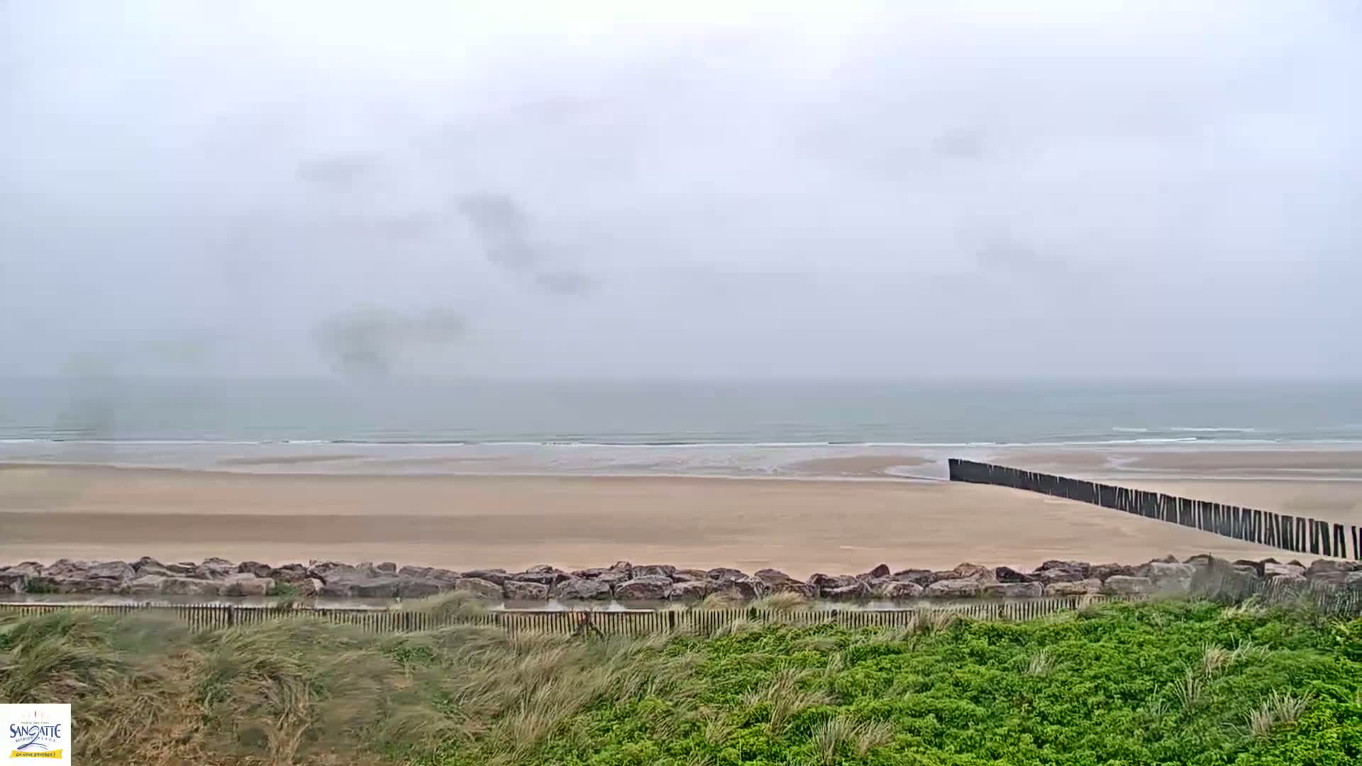 A wide sandy beach stretches to a calm, gray sea under an overcast sky, partially protected by a long, dark barrier.