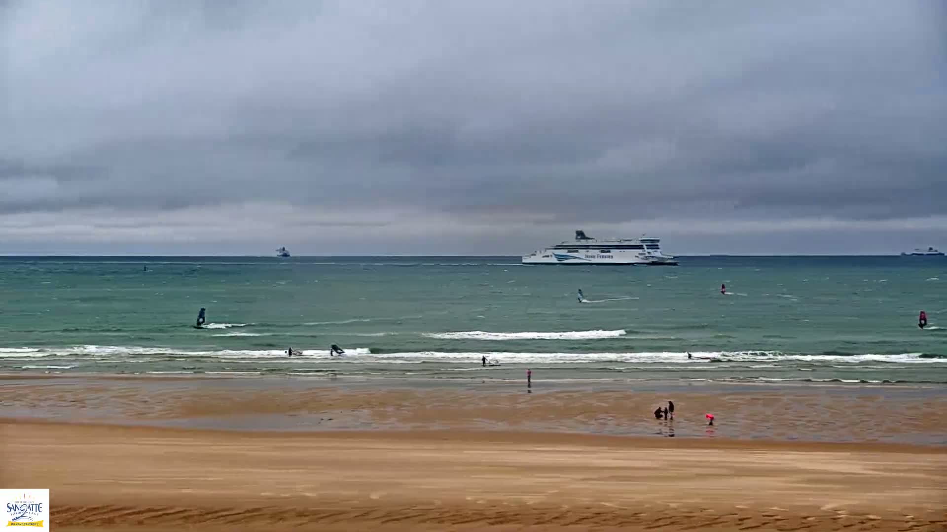 A sandy beach with several people and windsurfers in the water, a ferry in the distance, under an overcast sky.