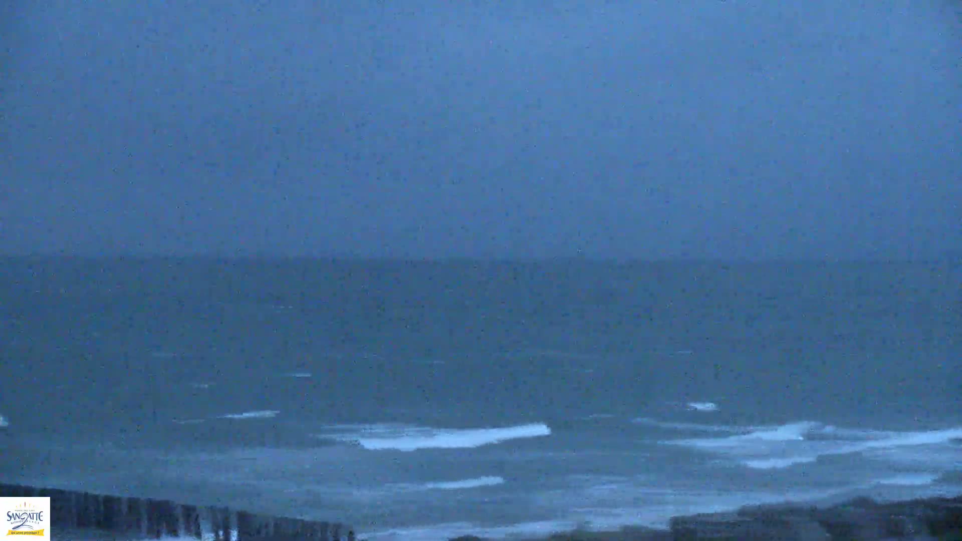 A dark, stormy ocean with small waves is visible from a high vantage point, showing a seawall in the bottom foreground.