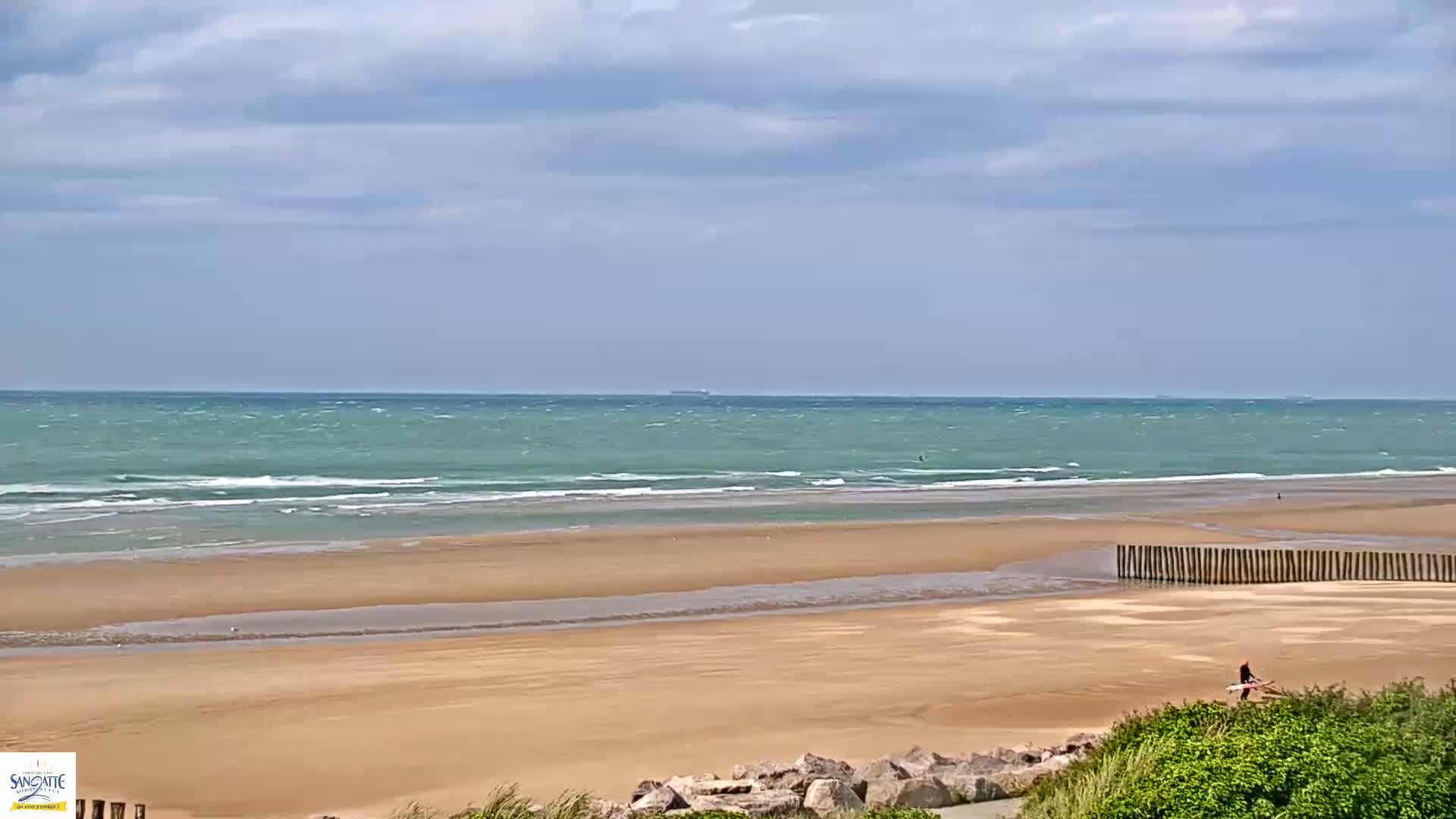 A wide sandy beach with a person carrying a surfboard is seen under a partly cloudy sky, with a gently wavy ocean in the background.