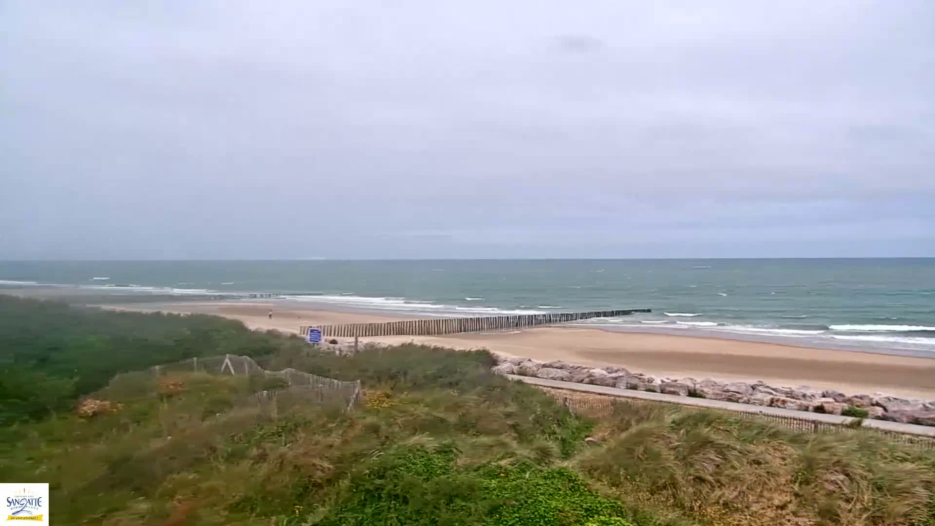 A sandy beach with a wooden wave break stretches along a windy, gray-sky ocean under an overcast sky.