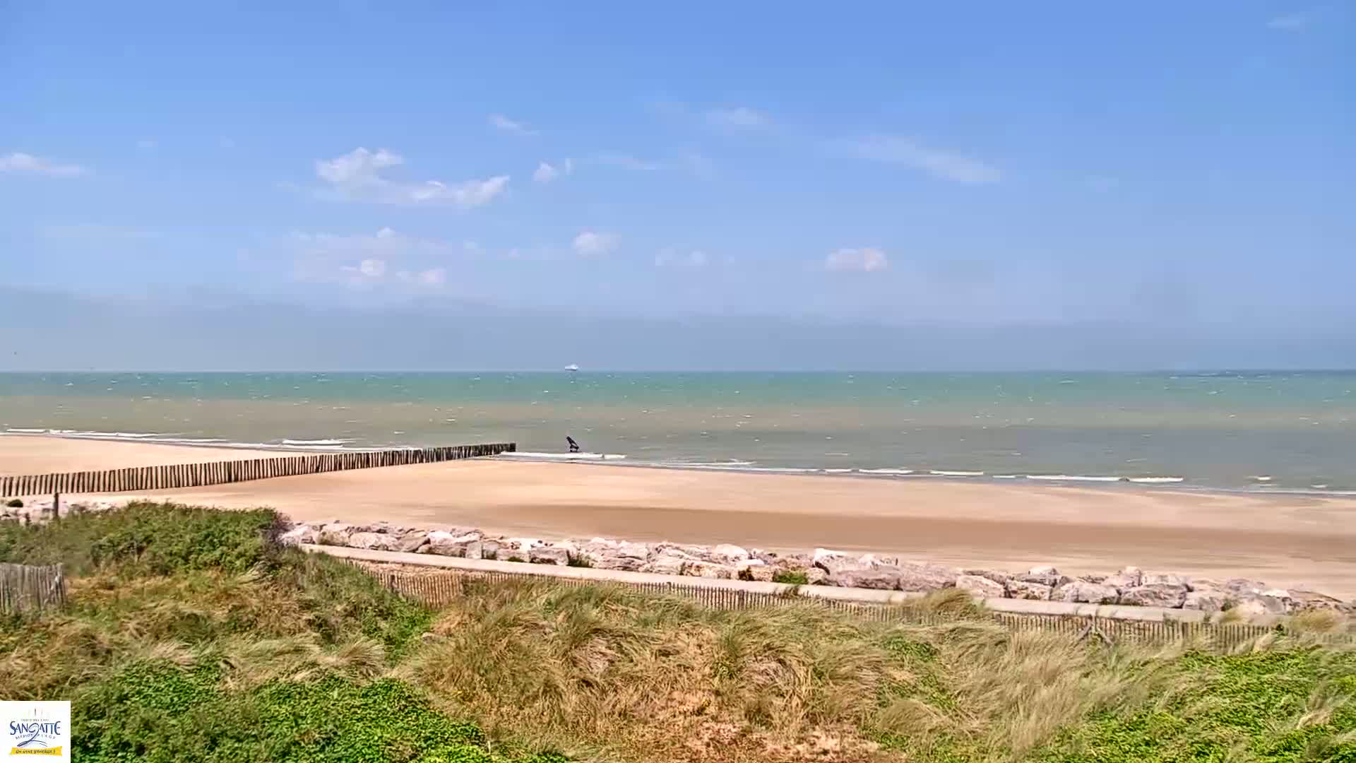 A sandy beach with a wooden fence and a calm, light-green sea under a mostly sunny sky.
