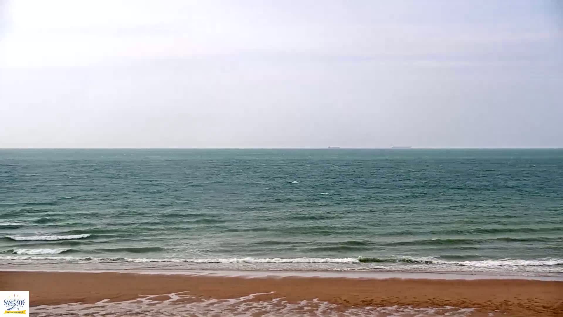 A tan sandy beach meets a teal sea with small waves under an overcast sky.