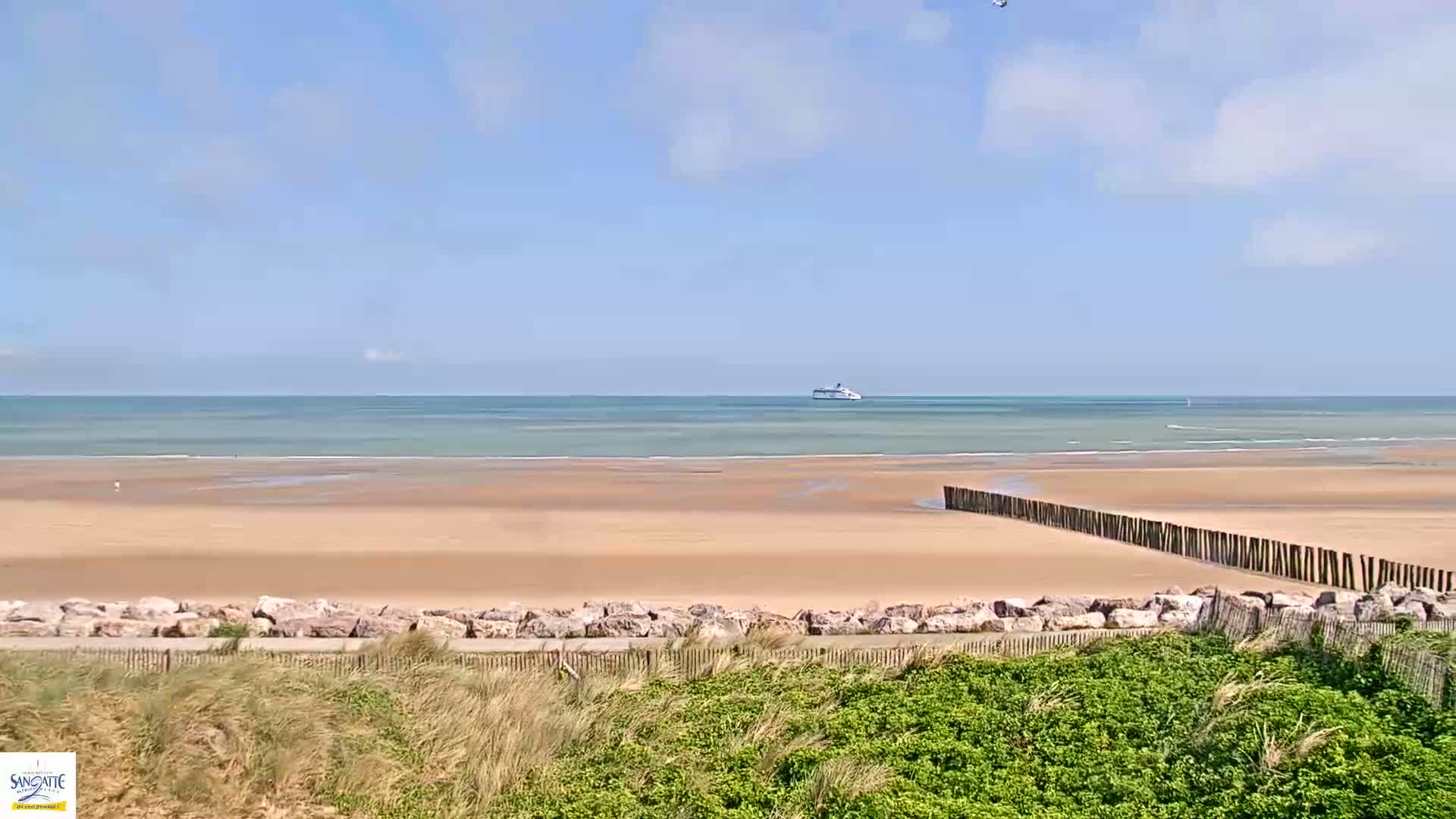 A sandy beach stretches to a calm ocean under a mostly clear blue sky, with a large ship visible in the distance and a wooden barrier partially separating the sand from the grassy dunes.