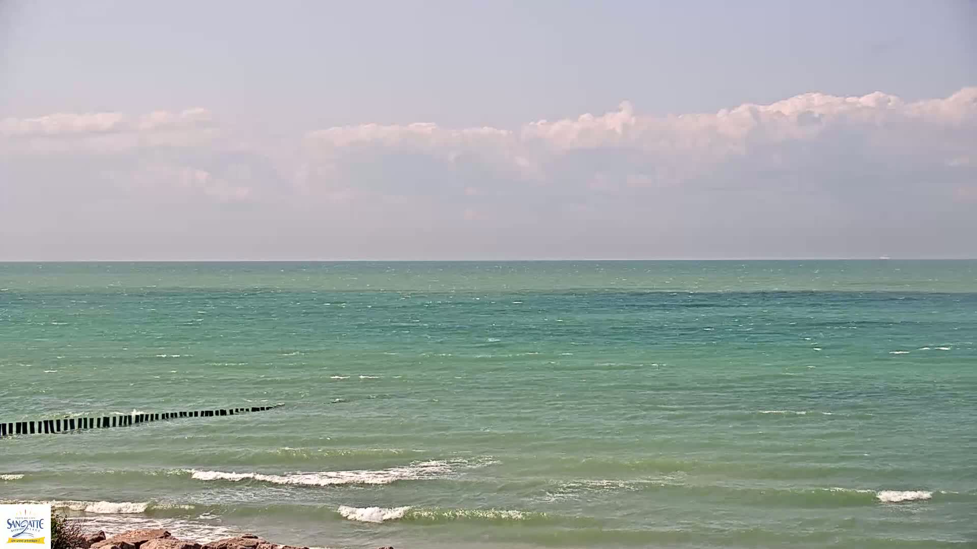 A mostly sunny day shows a calm, teal-green ocean with small waves lapping a rocky shore, partially protected by a breakwater.
