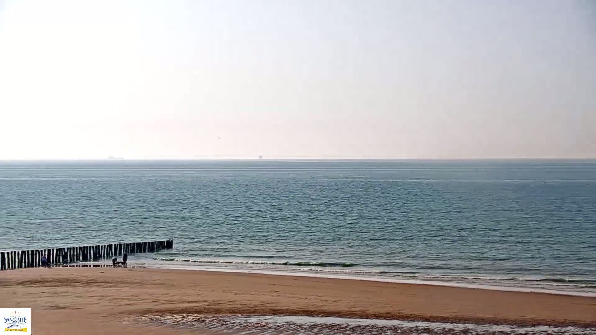 A sandy beach meets a calm, teal sea under a hazy sky, with a few people near a low wooden breakwater.