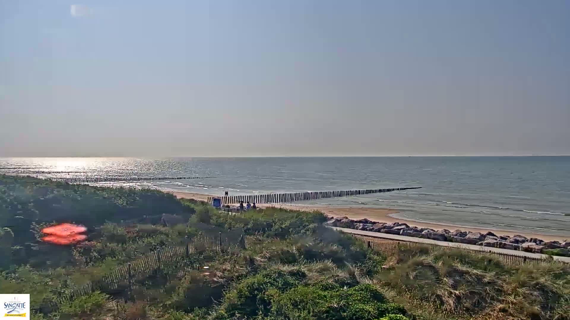 A sandy beach with a calm ocean under a clear sky features a long, low wooden structure parallel to the shoreline, with a few people visible near the water's edge.