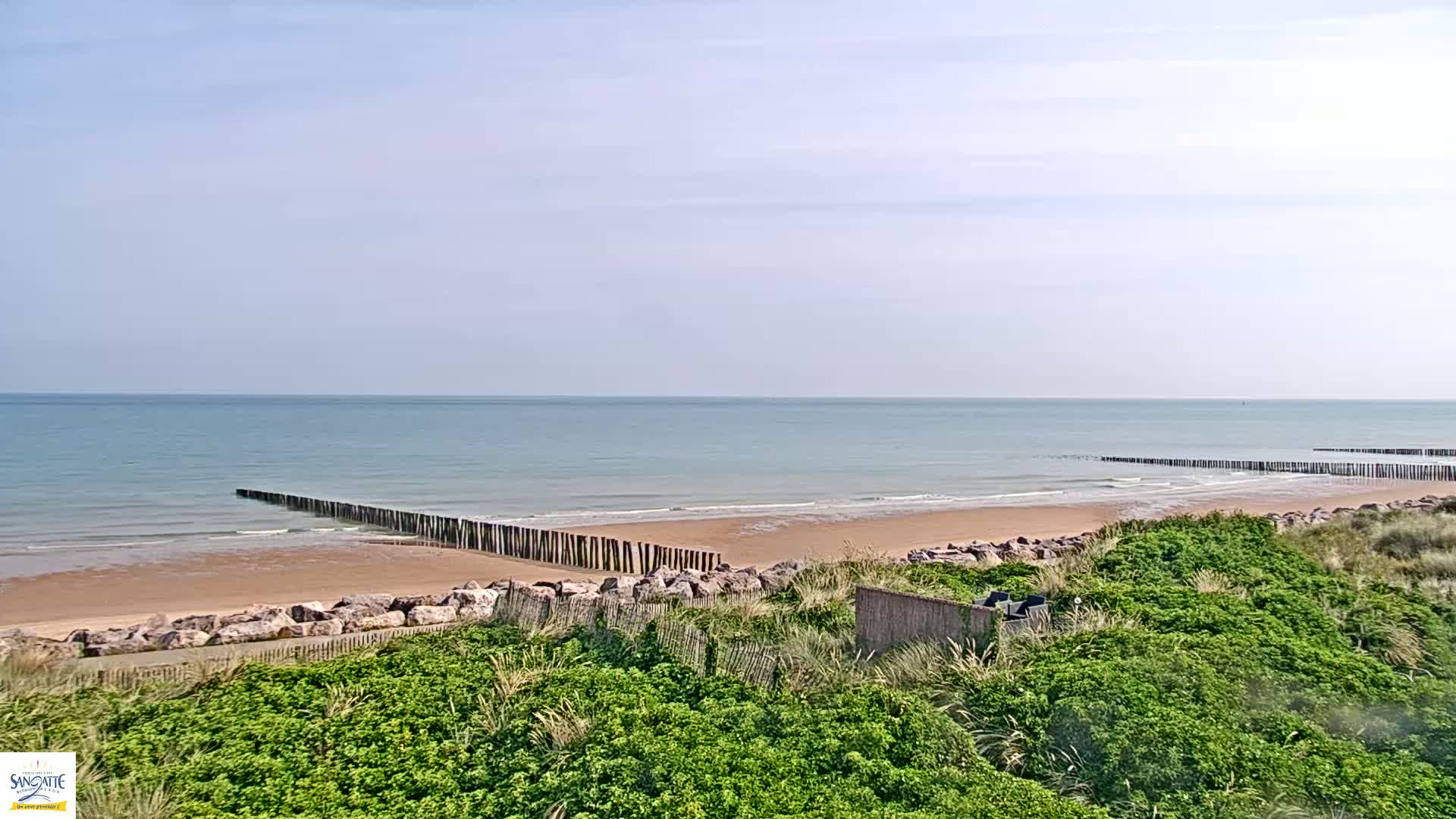 A sandy beach with calm ocean waves and wooden barriers stretches out under a mostly clear sky.