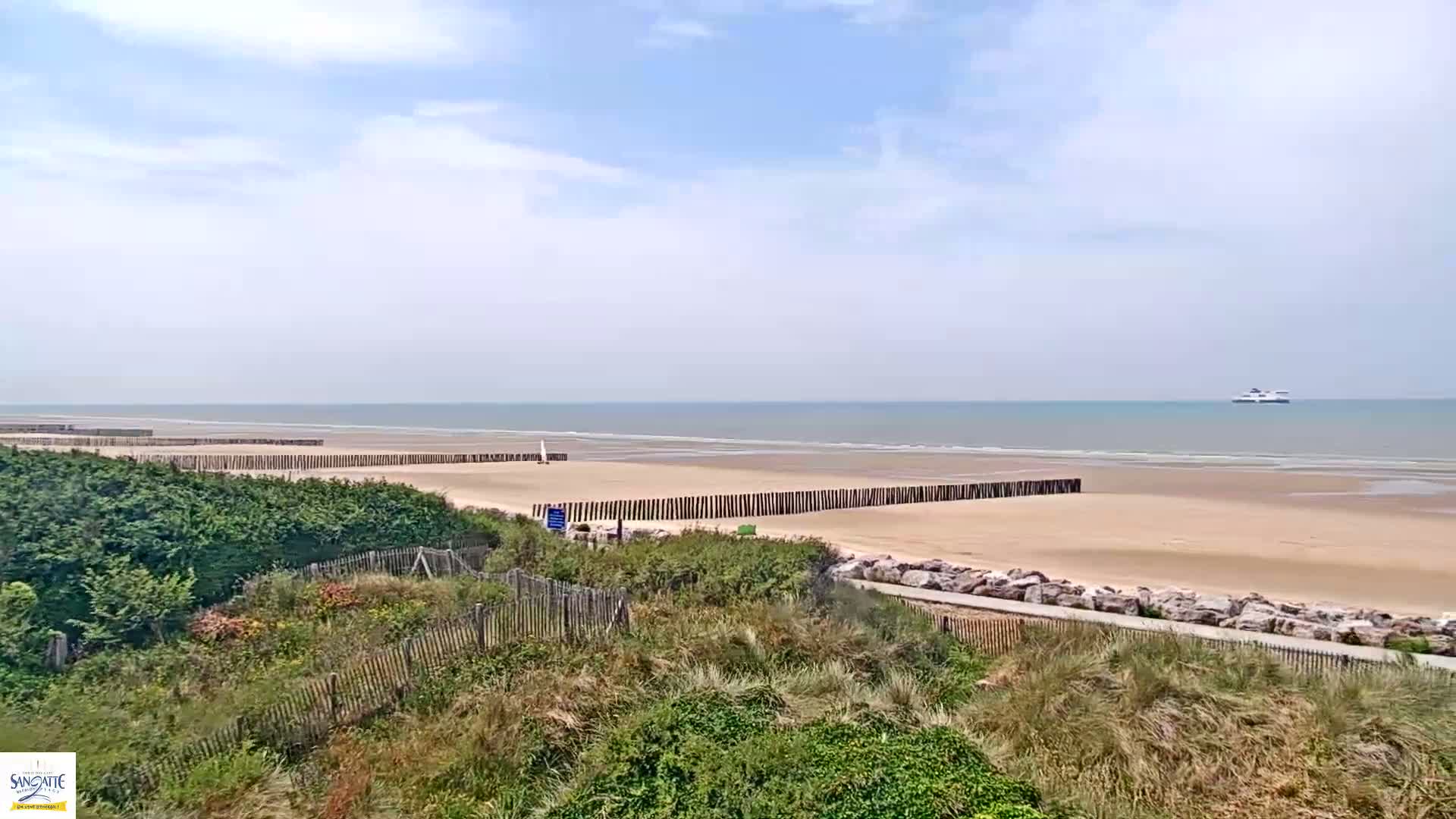 A sandy beach with wooden breakwaters stretches along a calm ocean under a partly cloudy sky, viewed from a dune covered in vegetation.
