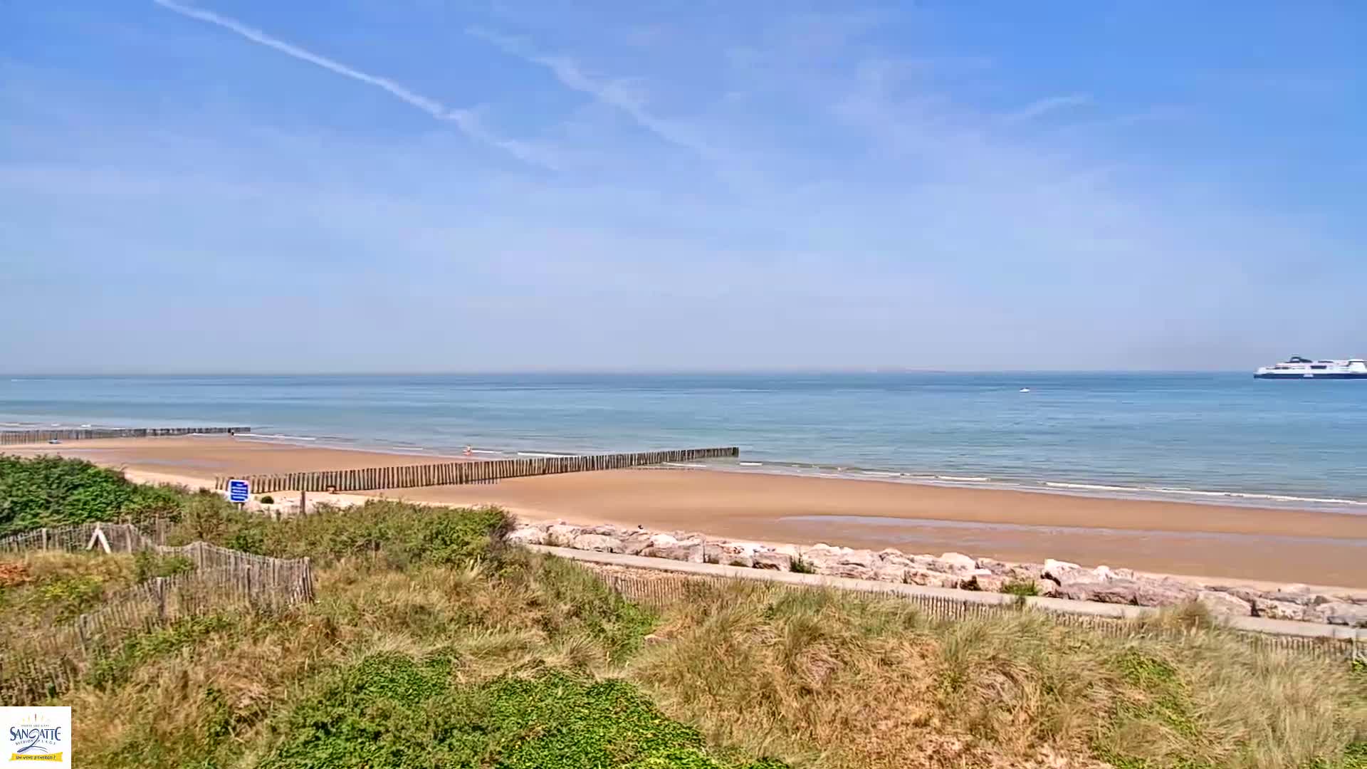 A sandy beach with calm ocean waves under a clear blue sky features a distant ferry and wooden barriers along the shore.