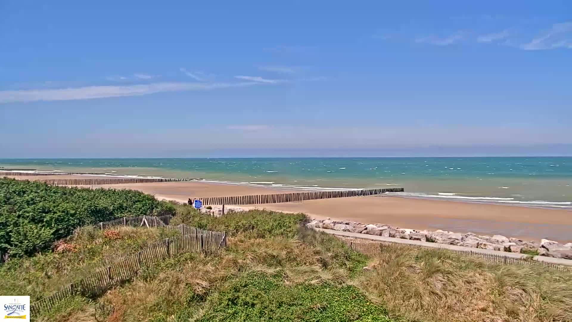 A sandy beach with a wooden fence stretches along a teal ocean under a mostly clear blue sky with a few wispy clouds.