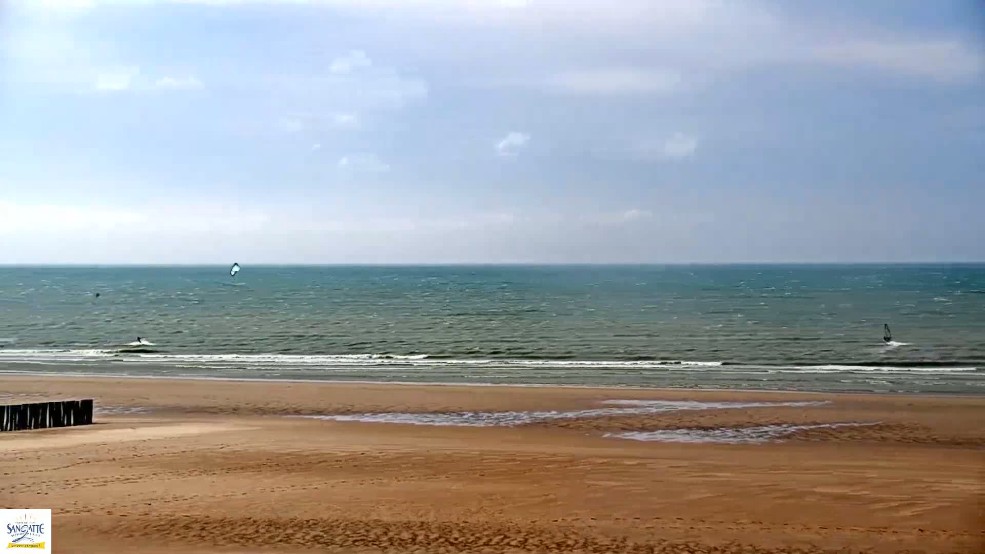 A wide sandy beach with a few windsurfers visible on a somewhat windy, partly cloudy day.