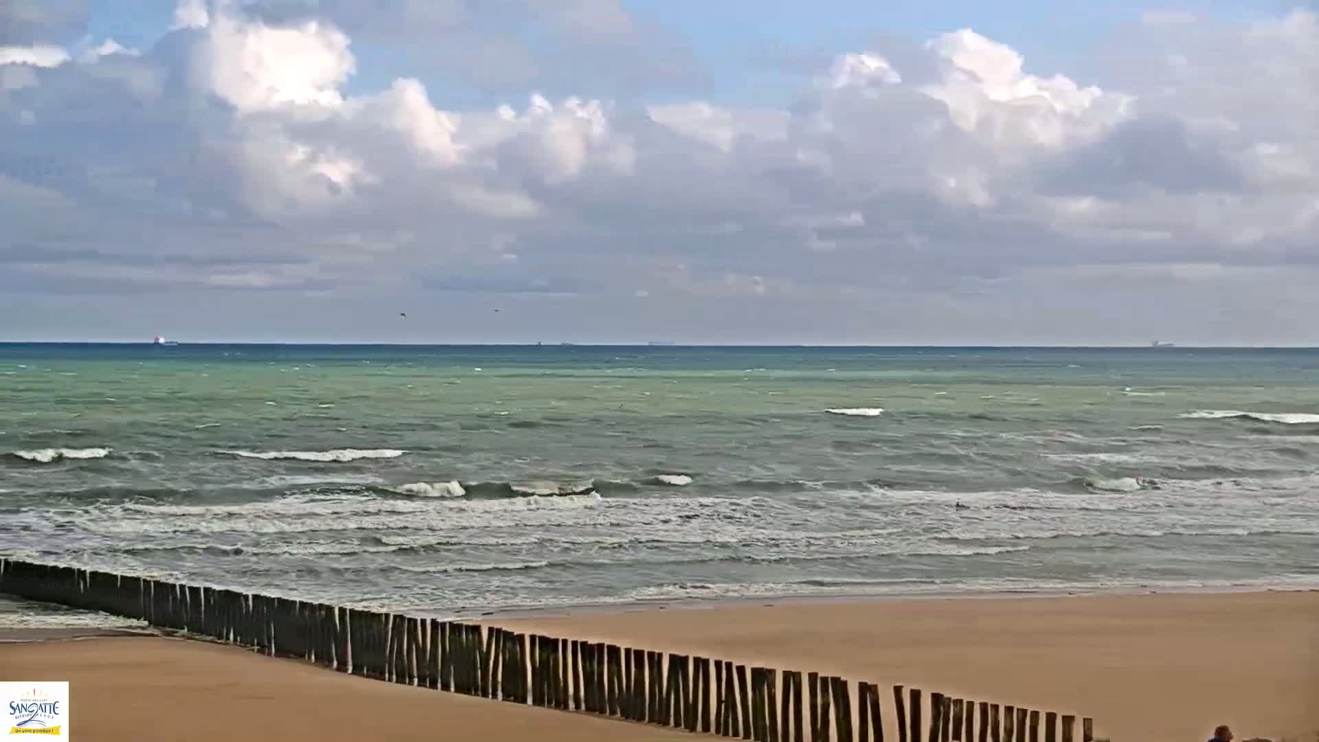 A sandy beach with a wooden fence separating it from a choppy sea under a partly cloudy sky.